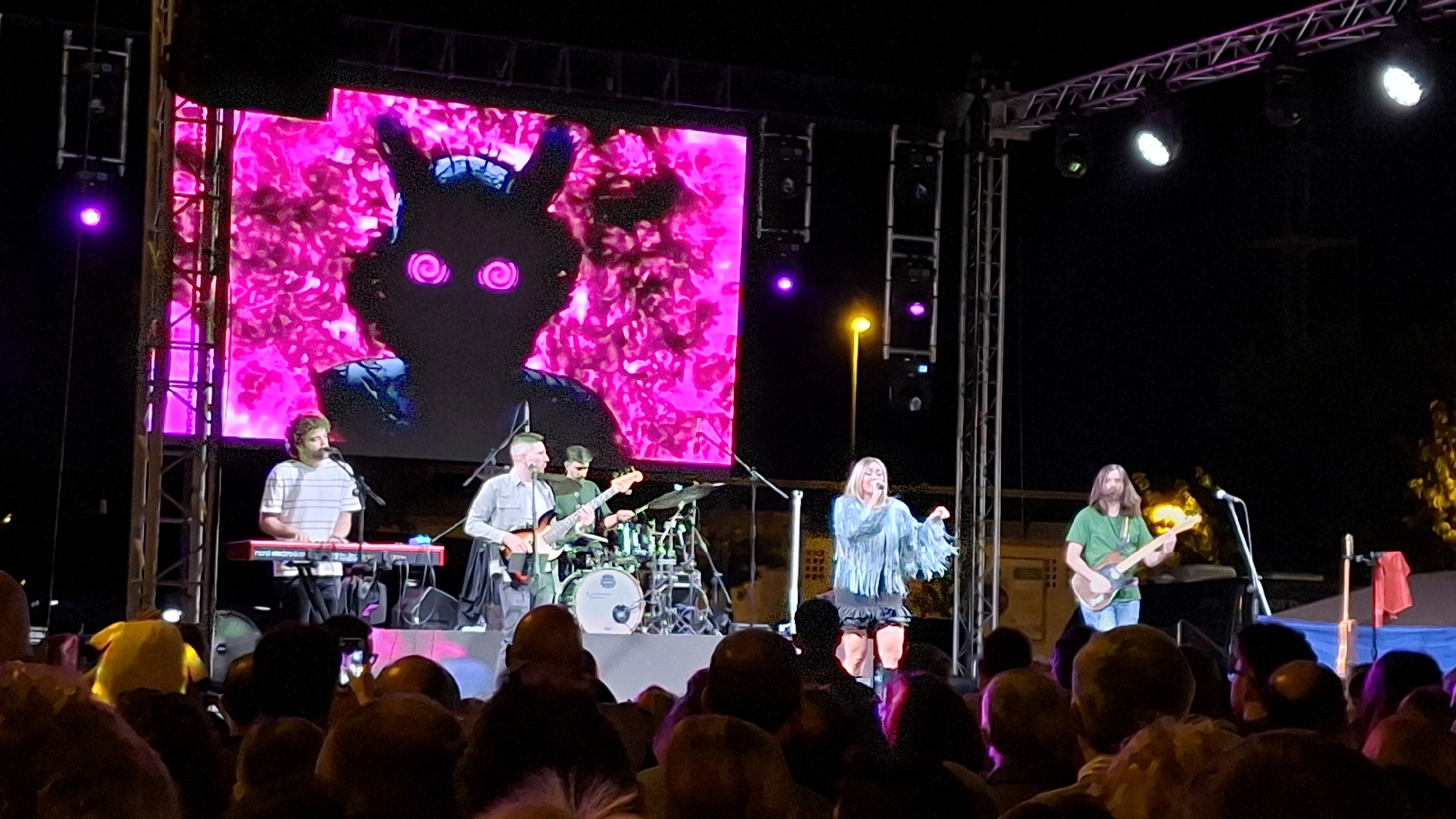 The image captures an outdoor music performance at night in Alcañiz, Spain. A large LED screen behind the band displays a psychedelic, neon pink background with a stylized, shadowy figure featuring glowing purple eyes. On stage, a band performs in front of the screen. To the left, a musician plays a red keyboard and sings into a microphone. Next to him, another band member plays a guitar and sings. In the center background, a drummer is visible behind his kit. To the right of the center, a female vocalist with a fringe-detailed blue top and dark shorts sings into a microphone. Further to the right, a male guitarist in a green t-shirt plays a guitar. A crowd of people is gathered in the foreground, watching the performance, with some holding up their phones to capture the event. Stage lighting and structural elements of the concert setup are visible.