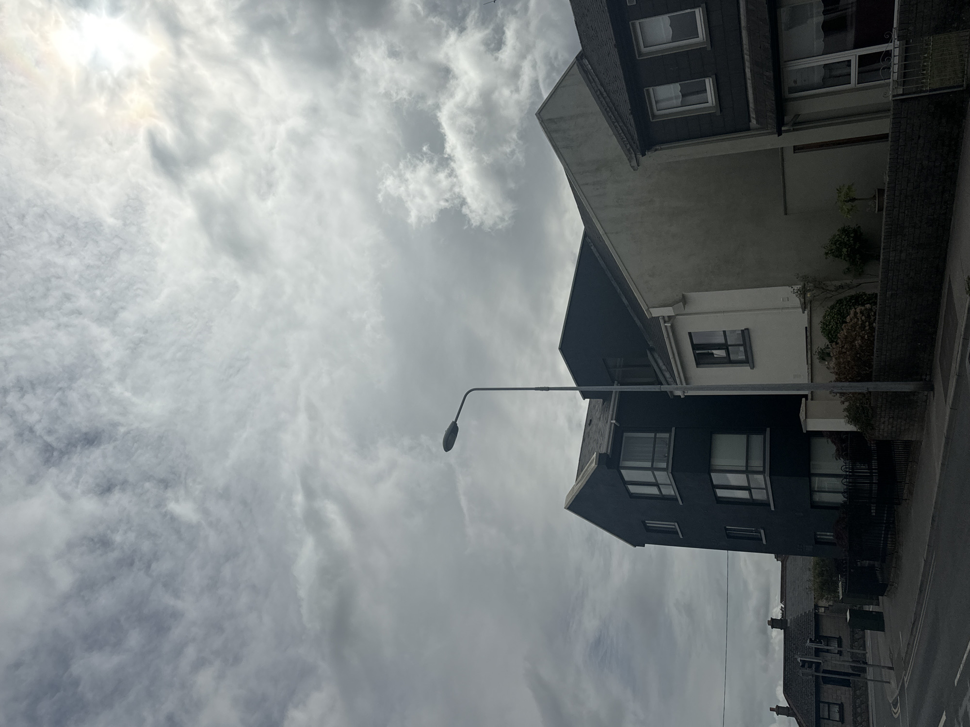 The image captures a street-level view of buildings under a cloudy sky in Waterford, Ireland. A dark, modern building with large windows stands prominently on the left, adjacent to a lighter-colored, older building. To the right, a dark-roofed house with white-framed windows is visible. A street lamp with a modern design is positioned between the buildings, near the center of the frame. The sky is filled with dramatic, textured clouds, with a bright, sunlit area visible in the upper right corner, suggesting the sun is partially obscured. In the background, more houses with traditional pitched roofs and chimneys can be seen. The foreground shows a paved road and sidewalk, with low brick walls and some vegetation. The overall impression is of an urban or suburban street scene on a day with varied weather conditions.