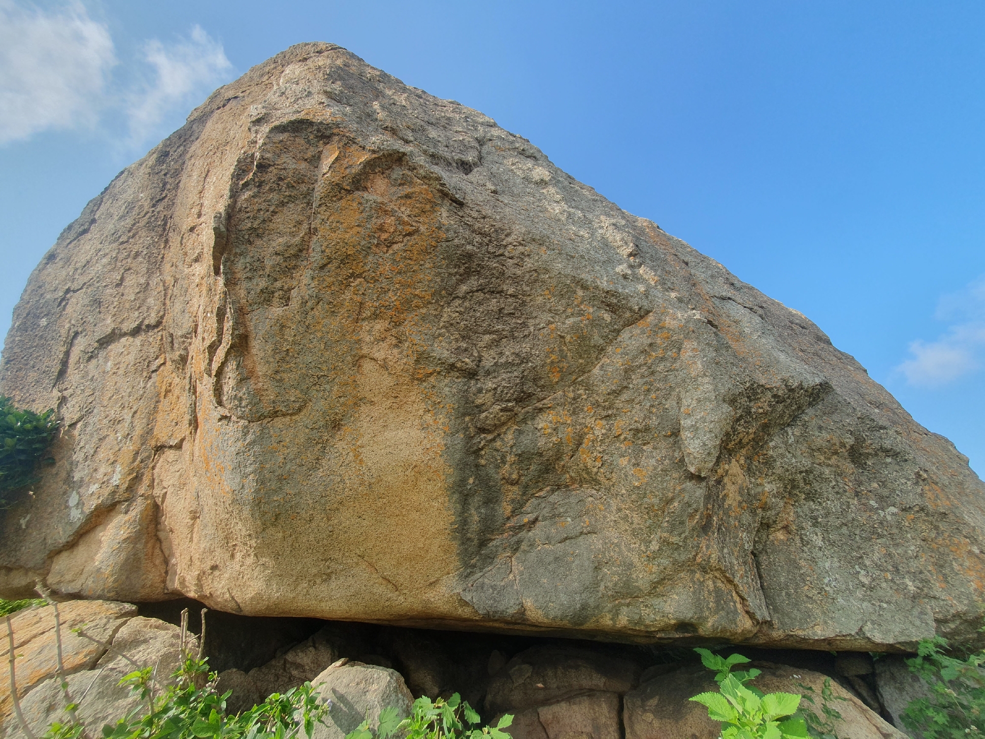 A large, weathered boulder, tinged with orange and grey, dominates the foreground of this outdoor scene, positioned at the foot of Gura Top, Nigeria. The boulder's rough surface displays numerous cracks and fissures, indicative of long-term exposure to the elements. It rests on a base of smaller rocks, creating a sheltered space beneath its overhang. Lush green vegetation sprouts from the ground around the boulder, adding a touch of vibrant life to the rocky landscape. The sky above is a clear, brilliant blue, with a few wispy white clouds drifting lazily across it, suggesting fair weather. The overall impression is one of natural grandeur and rugged beauty.
