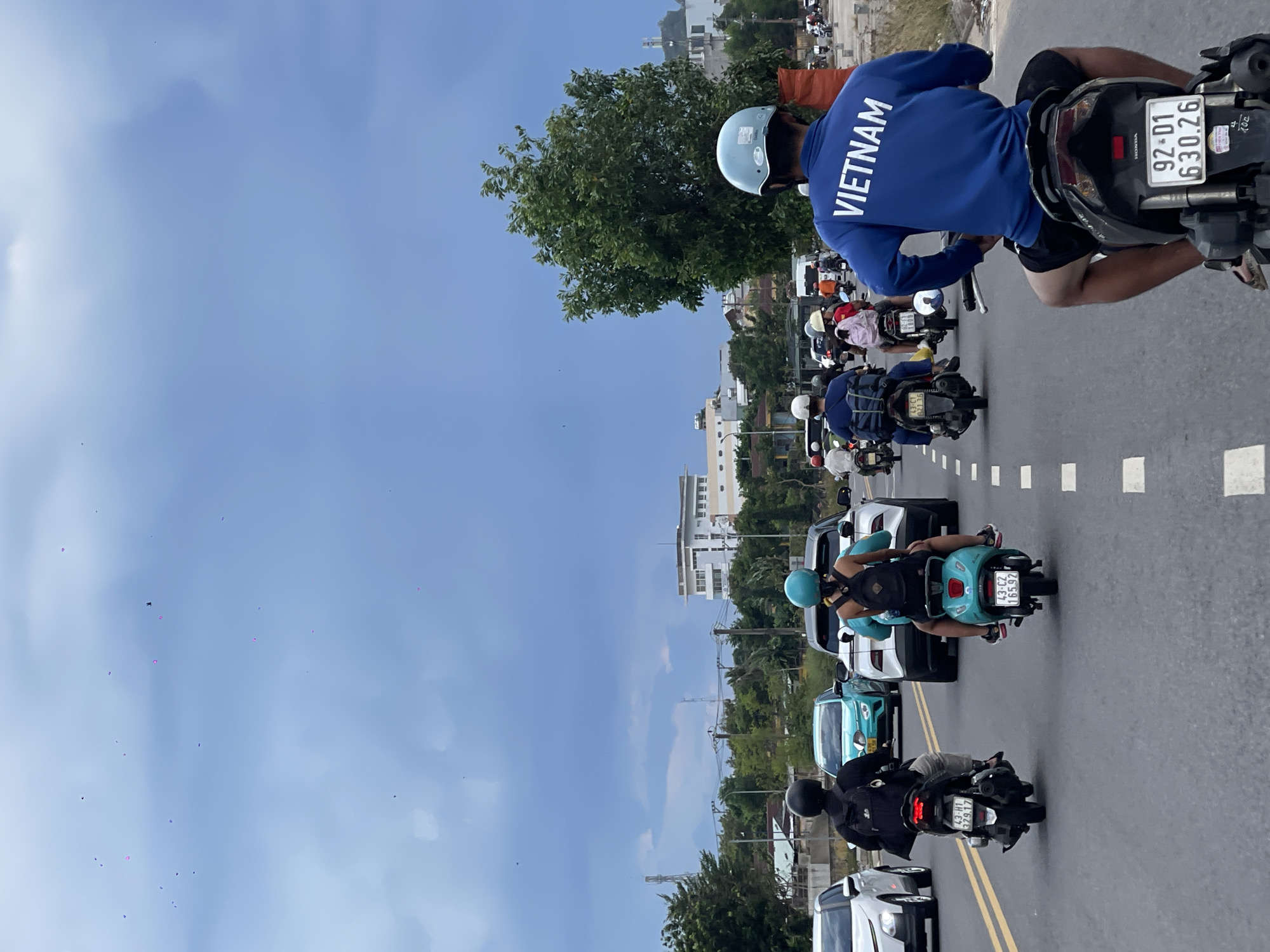 A group of people are riding motorbikes on a paved road in Đà Nẵng, Vietnam. The sky is a bright blue with some white clouds, suggesting it is daytime. The road is lined with trees and buildings, indicating an urban or suburban setting. The motorbikes are moving in the same direction, with several people wearing helmets for safety. One person in the foreground wears a blue sweatshirt with "VIETNAM" printed on the back in white letters. Another rider wears a teal helmet and a backpack. The overall scene appears to be a typical day of commuting or travel in Vietnam. The license plates of the vehicles are visible, one reading "92-D1 630.26". Another license plate reads "43-CZ 165.97".