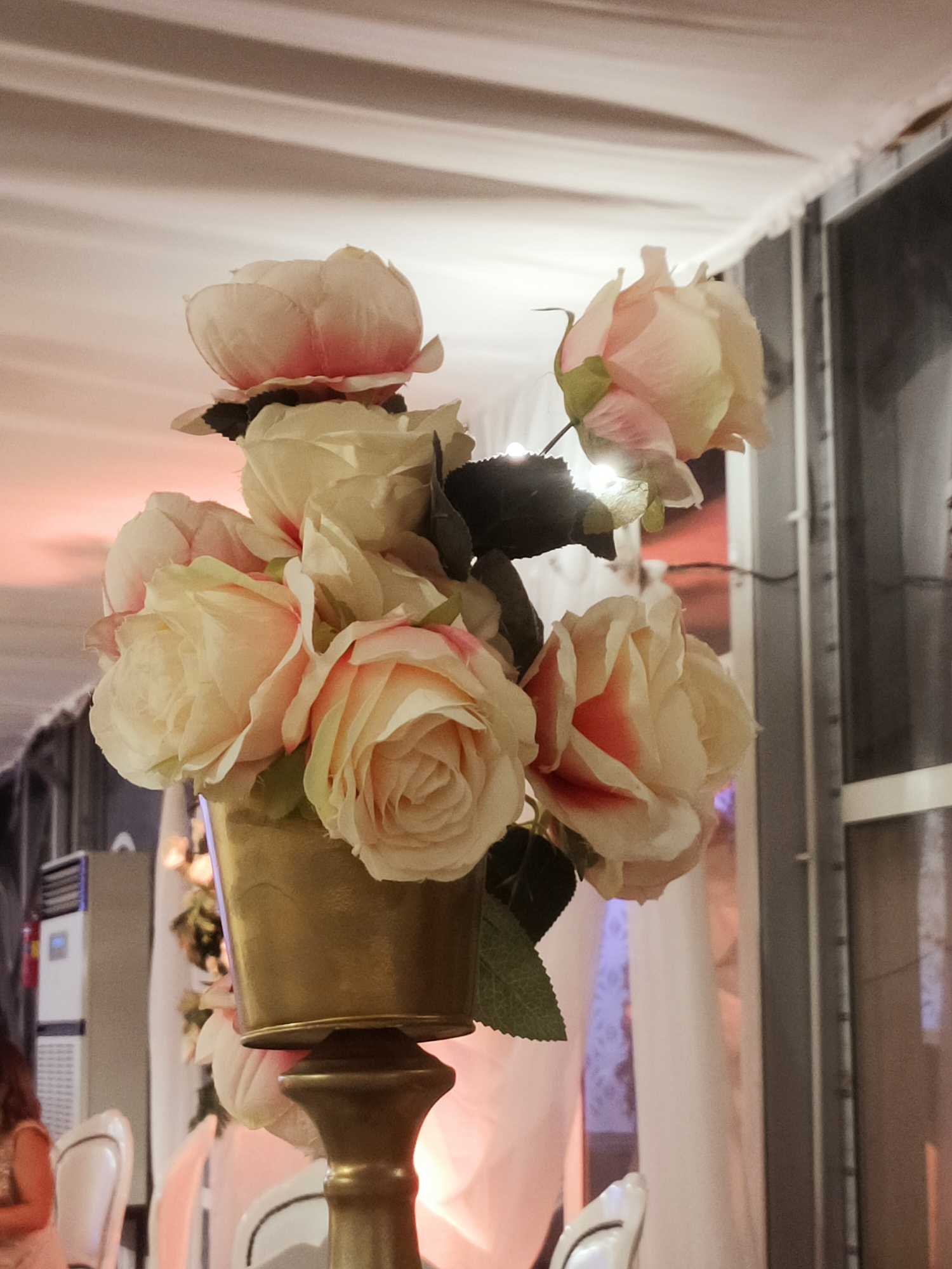 A close-up shot focuses on a gold vase holding a bouquet of artificial roses. The roses are predominantly cream-colored with hints of pink and red on their petals. The vase is cylindrical with a flared rim and sits atop a gold pedestal. In the background, soft, diffused light suggests an indoor setting, possibly a banquet hall or event space. Faintly visible are white chairs and draped fabric, indicating a formal or celebratory occasion. The overall impression is one of decoration and ambiance. The location of this scene is not discernible from the image itself.