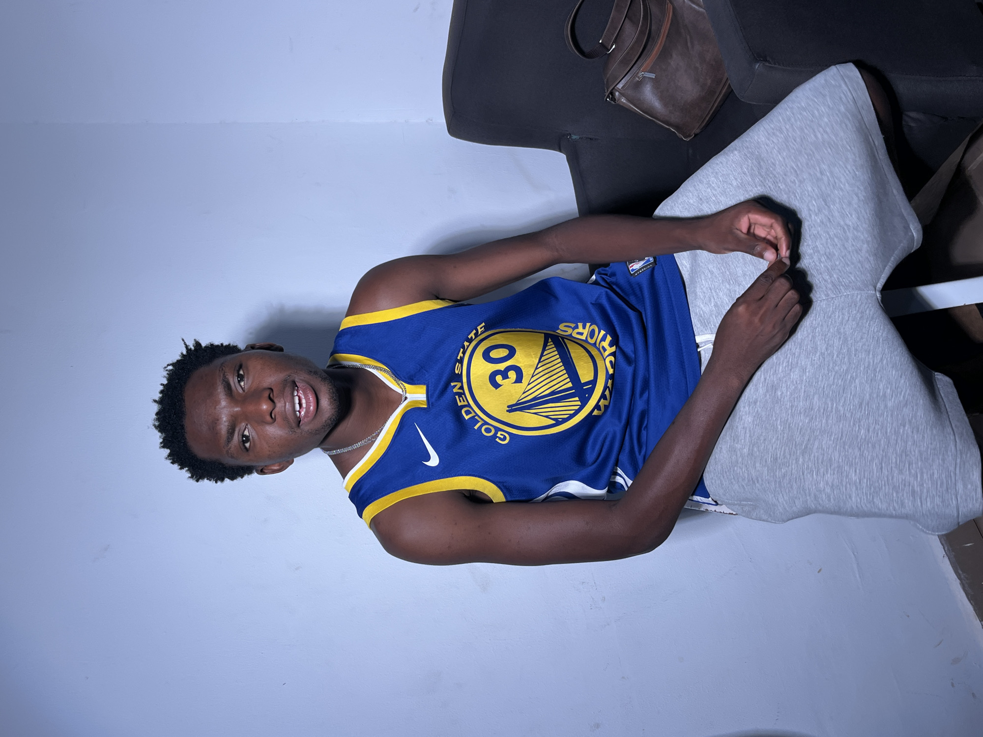 A young Black man wearing a blue and yellow Golden State Warriors basketball jersey with the number 30 on it is lying down, with his head propped up on a large grey pillow. The jersey has the Nike logo and the words "GOLDEN STATE WARRIORS" visible. He is wearing a silver chain. He is looking at the camera with a slight smile, and his mouth is open. His hair is short and curly.
The man is lying on what appears to be a dark couch, with a brown bag visible on the backrest. The background is a plain, light-colored wall. The lighting suggests it is indoors, possibly in a living room or studio setting, with a soft light illuminating the man and the immediate surroundings. The image is taken from a slightly high angle, looking down at the man. There are no obvious indications of the time of day or weather conditions. The overall mood of the image is relaxed and casual. The location is unknown.
