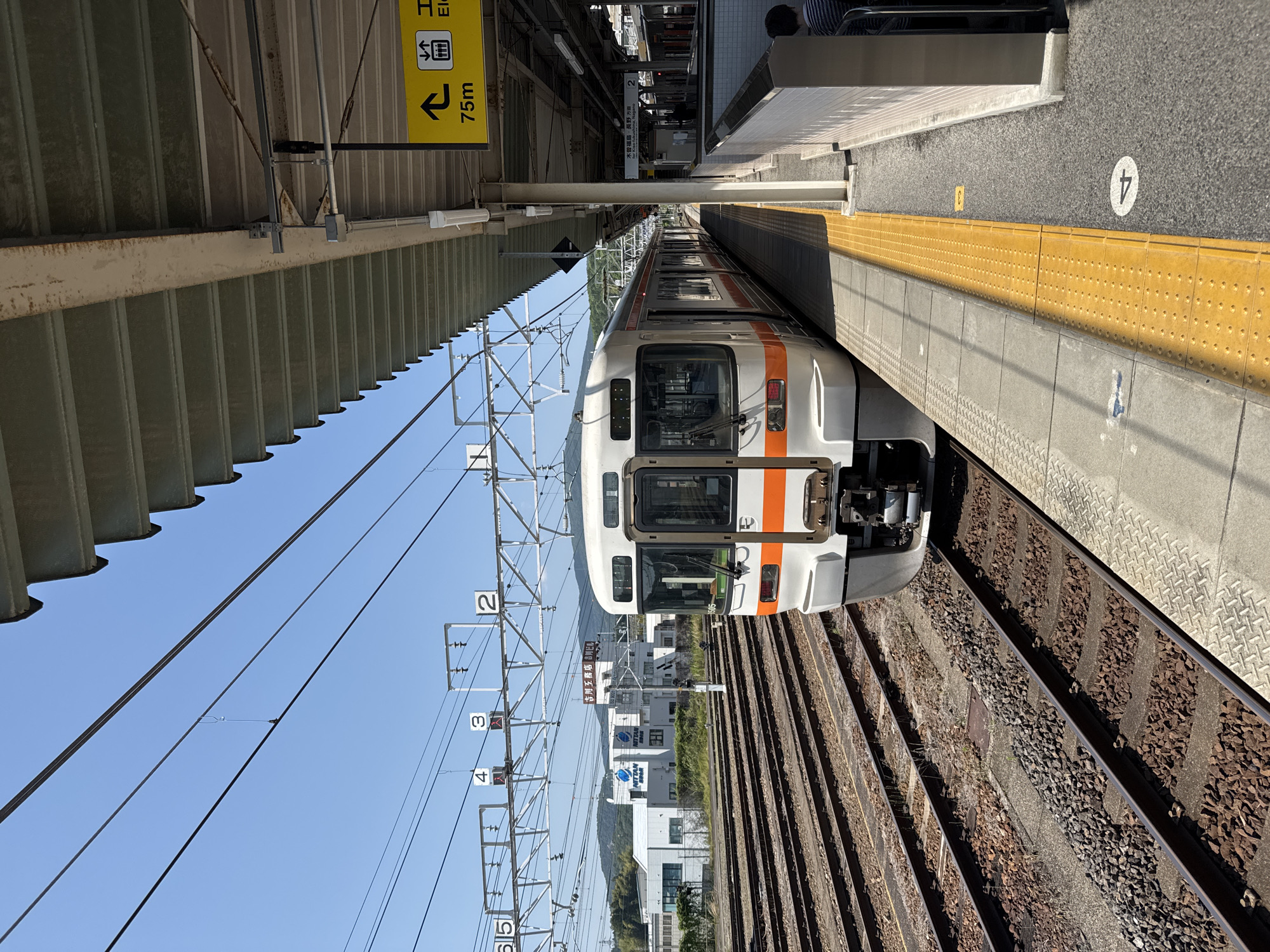 The image shows a train at a station platform. The train is white with an orange stripe, and the station platform has a yellow tactile strip along the edge. A sign indicates that elevators are 75 meters in the indicated direction. A sign has the text "吉川工務店 吉川ビル" (Yoshikawa Construction Yoshikawa Building). Overhead electrical lines are visible above the train tracks, and the tracks are lined with ballast stones. Buildings can be seen in the distance behind the train. The platform is marked with the number "4". The location appears to be Nakatsugawa, Japan based on the text of the sign.