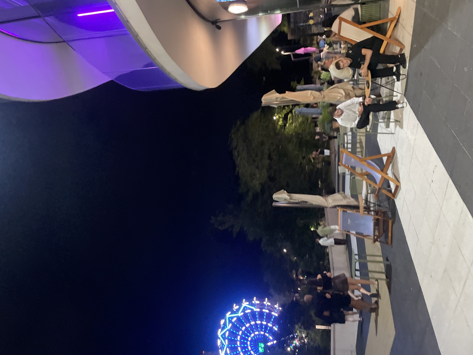 The image captures an outdoor scene at night, likely in a city, featuring a Ferris wheel in the background to the left, illuminated with blue and white lights. In the foreground, a modern building with a curved facade and purple lighting is visible on the right. Several people are present in the scene, some seated at outdoor tables and others walking. Two men are seated at a small table in the foreground, one looking at his phone. Other individuals are seen in the mid-ground, some walking and others seated in lounge chairs. Umbrellas and outdoor furniture are scattered throughout the area. The ground is paved with a pattern of dark and light tiles. The overall atmosphere suggests a social gathering or entertainment venue at night.