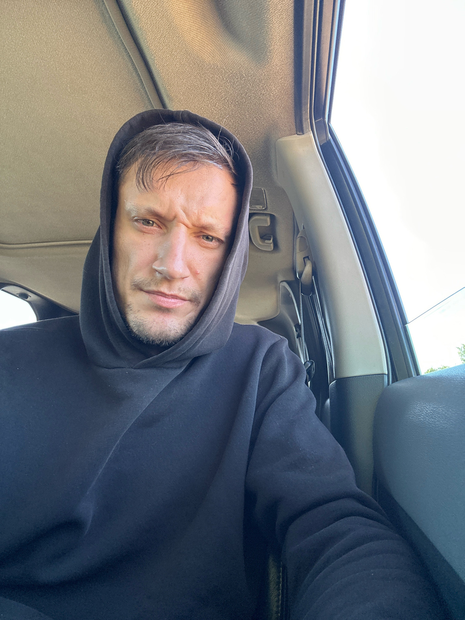 The image is a selfie taken from inside a car, likely during the daytime. A man with short, graying hair is wearing a black hoodie, which he has pulled up over his head. His eyebrows are furrowed, and he appears to be looking directly at the camera with a serious or concerned expression. The interior of the car is visible, including the headliner and part of the car door and seat. The lighting suggests an outdoor environment, possibly a road or residential area. There is no specific indication of the location being the city of Makiv, Ukraine.