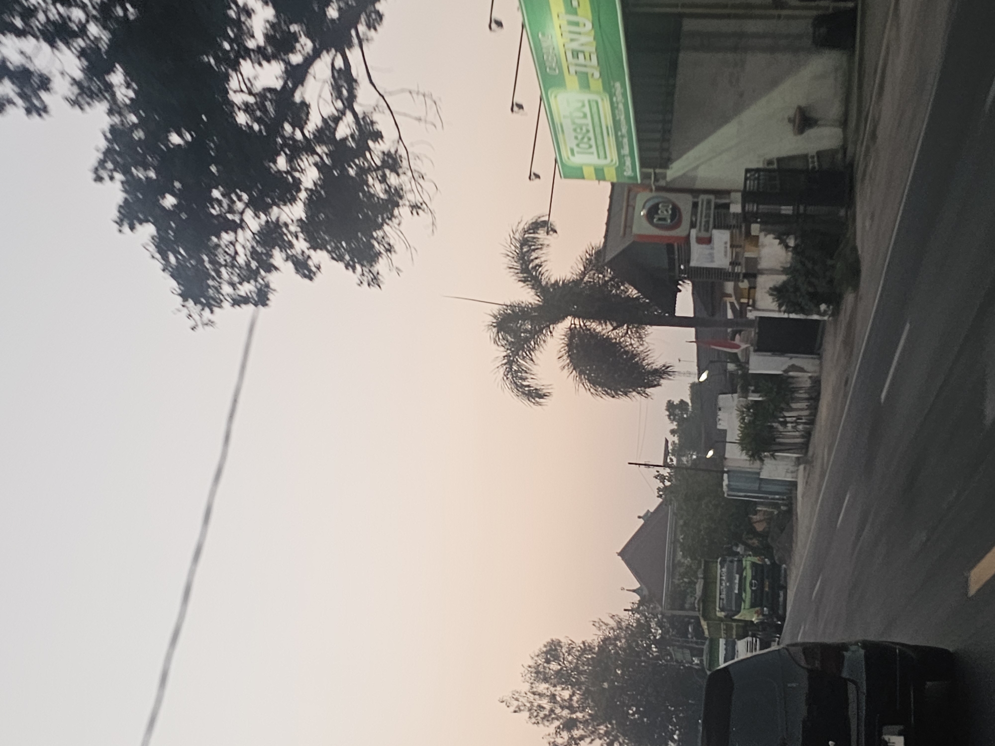 The image depicts a street scene in Jenu, Indonesia, at what appears to be dusk or dawn due to the soft, muted light and pale sky. A dark colored SUV is in the foreground, facing away from the camera and traveling on the right side of a two-lane road. A utility wire runs diagonally across the sky from the upper left to the middle of the image. To the right of the road, there is a green awning with white lettering that reads "Tosserbu JENU," indicating a store or business. A palm tree stands next to the awning. Further down the road, several trucks are parked. A building with a pitched roof and a dome-like structure on top, possibly a place of worship, is visible in the background. Trees and foliage are scattered throughout the scene. The overall impression is a typical roadside environment in a town or city.