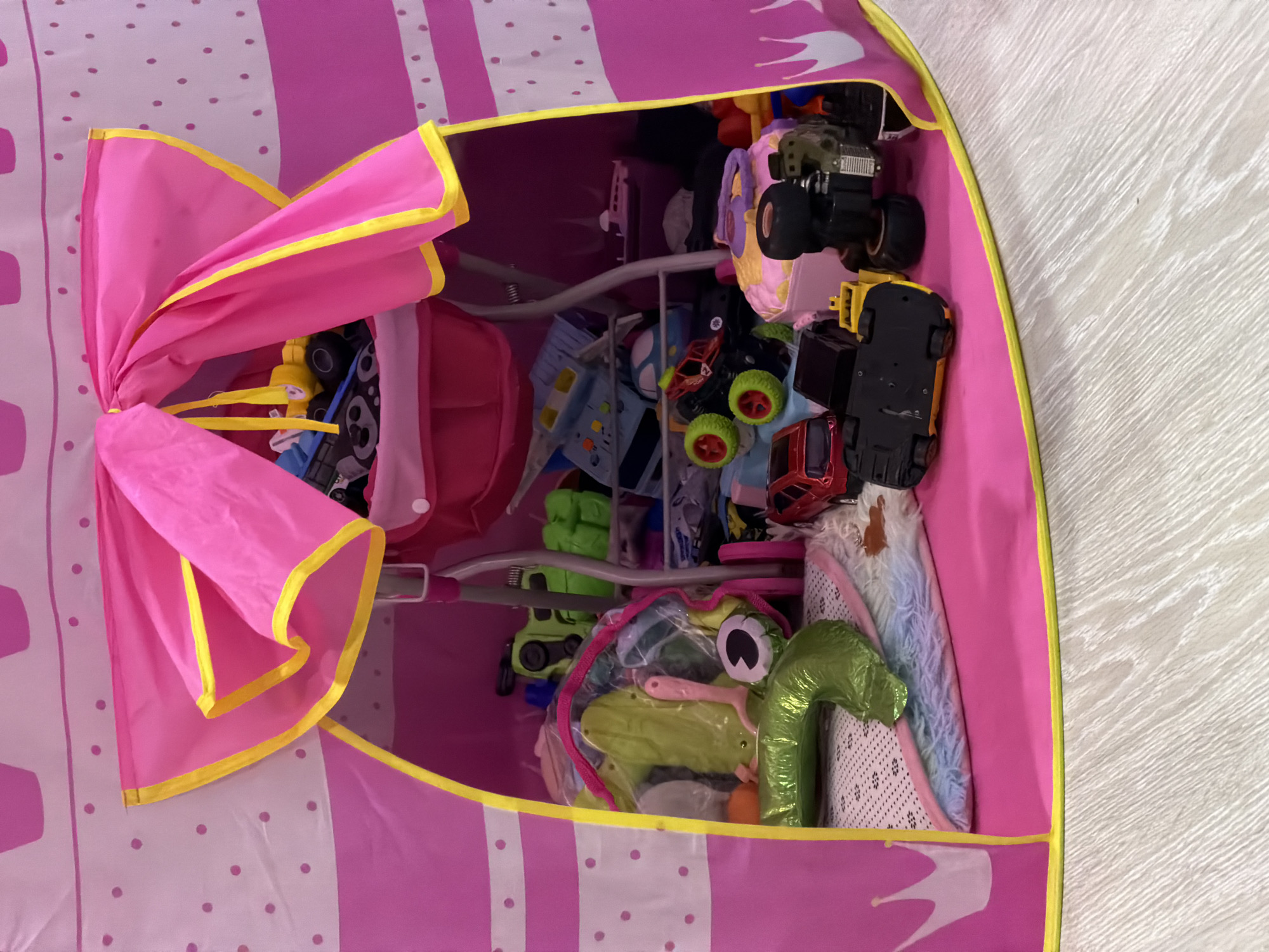 The image shows the inside of a pink toy storage unit, likely located in Noviy, Russia. The unit is divided into shelves and is filled with various toys, including toy cars, a green frog toy, and other unidentified items. The storage unit is made of fabric with pink and white stripes and polka dots, trimmed with yellow. The unit's door is tied open with a pink ribbon, allowing a full view of the contents. The floor beneath the unit is a light-colored, textured surface.