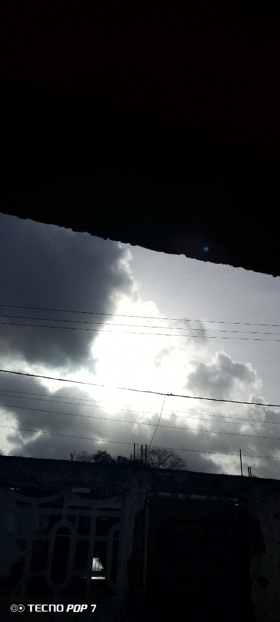 The image shows a section of the sky with dark, heavy clouds, some illuminated by a bright light source behind them, likely the sun. Multiple power lines cross the view. Below the sky, there's a dark, indistinct structure, possibly a building or wall, with hints of vegetation and fence details. The overall impression is of a potentially stormy or overcast day. The location, Altagracia, Venezuela, is indicated in the prompt. No people are visible in the frame. The word "Tecno Pop 7" is printed on the lower left corner of the image.