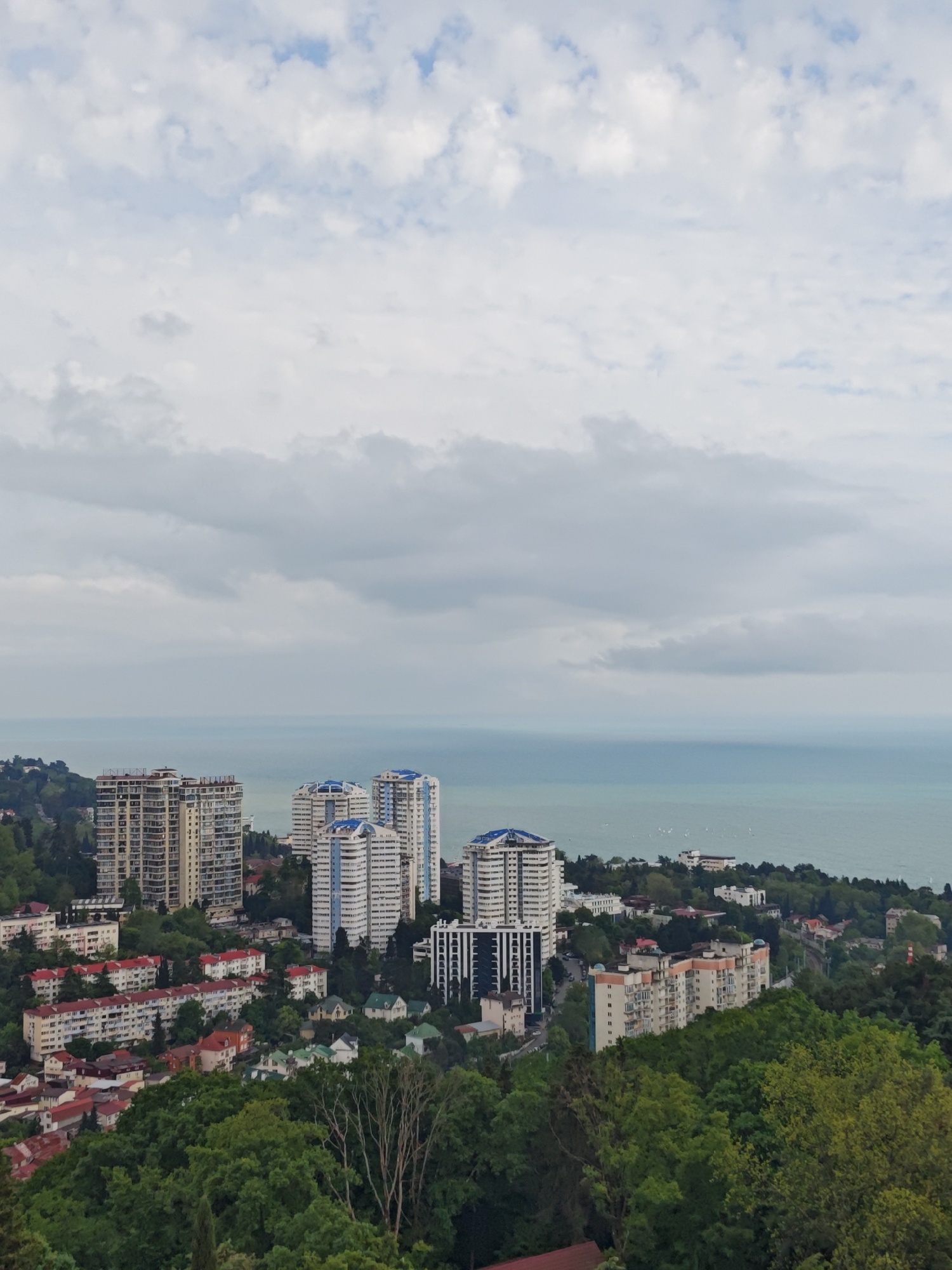 The image presents a panoramic view of Sochi, Russia, characterized by an urban landscape nestled along the coastline. Tall, modern residential buildings with distinct architectural features, including blue-tiled roofs, are interspersed with low-rise residential structures featuring red-tiled roofs. These buildings are situated amidst dense greenery, predominantly trees, which blend into the hilly terrain.
The Black Sea extends into the horizon, with faint hints of ships or boats visible in the distance. The sky is overcast with clouds, indicative of a diffused light, affecting the overall color palette of the scene. The composition of the image balances urban development with natural elements, typical of coastal cities. The buildings appear well-maintained, suggesting a functional urban environment.