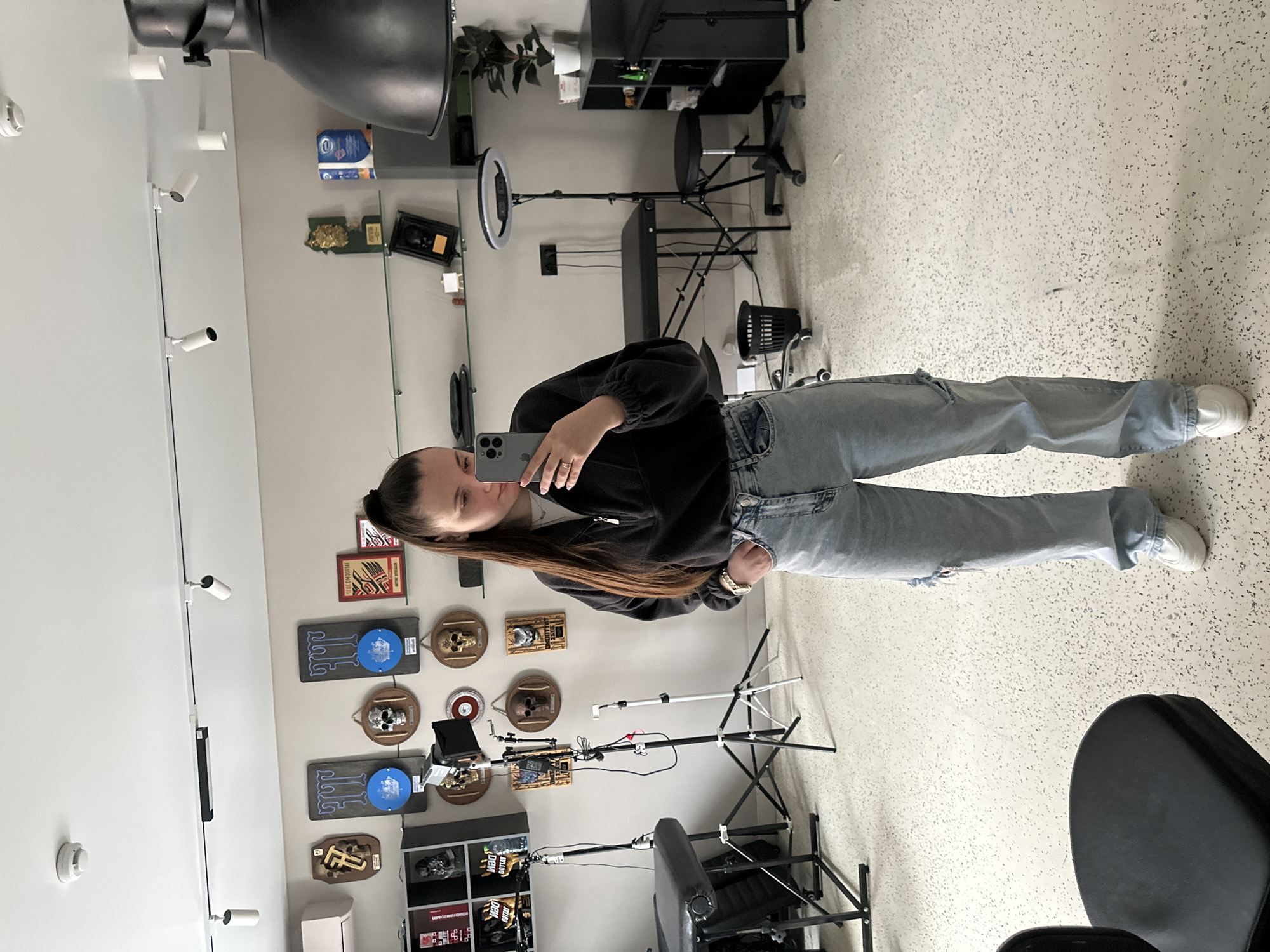 The image depicts a female taking a mirror selfie inside a tattoo studio in Rostov-on-Don, Russia. The individual, identified as a young adult female, stands centrally, with long dark hair in a high ponytail. She is dressed in a dark quarter-zip pullover, light blue ripped jeans with visible tearing on the left knee, and white athletic shoes. Her right hand holds a grey smartphone, angled to capture her reflection, while her left hand rests in her jeans pocket. A gold-toned watch is visible on her left wrist.
The studio features light grey walls and a light-colored, speckled floor. Two black adjustable tattoo chairs/beds are present, along with professional lighting equipment, including a softbox on a stand and a ring light on another stand. Wall decor comprises multiple skull-themed plaques, awards, and framed items. Visible Cyrillic text, notably "DEN TATTOO" on items located on a black shelving unit to the left, indicates the business type and Russian location. Overhead, a white ceiling houses track lighting and a smoke detector. Additional objects include a black adjustable wheeled stool, a black trash can, black shelving units containing supplies and decorative busts, an air conditioning unit on the upper left wall, and a potted plant on the right side.