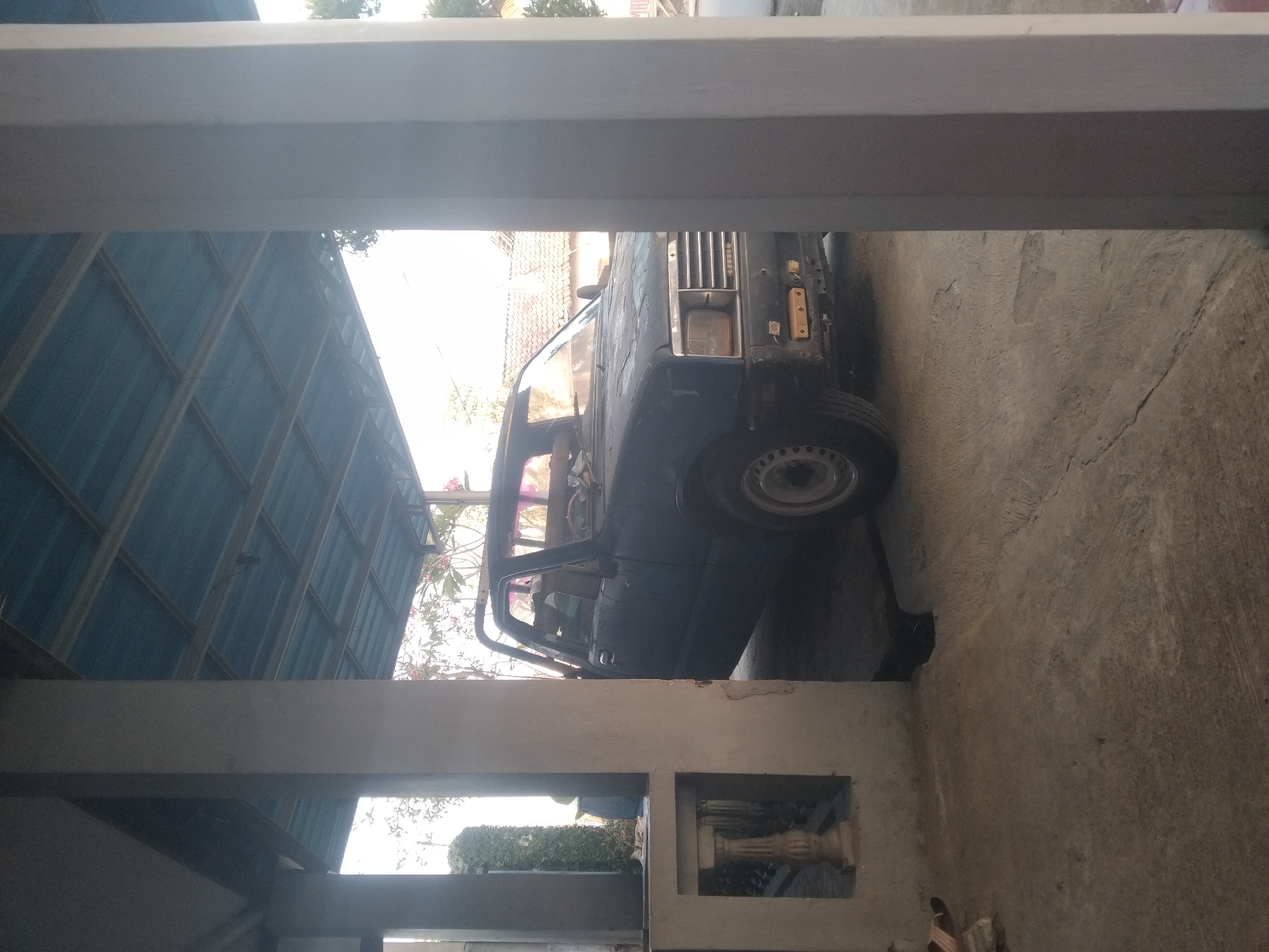 The image depicts a dark-colored pickup truck parked under a blue corrugated metal roof supported by concrete pillars. The truck is positioned in what appears to be an open-air carport or garage. The front of the truck, including its grille and headlights, is visible. The ground is concrete and shows signs of wear and cracking. In the background, foliage and rooftops of houses are visible, suggesting an urban or suburban environment. To the left of the truck, a portion of a decorative concrete structure with an ornamental pillar is visible. The scene is illuminated by natural light, casting shadows and highlighting the textures of the concrete and metal surfaces. The context of Tempeh, Indonesia is provided, suggesting the geographical location of this scene. There are no people clearly visible interacting with the truck or the environment.