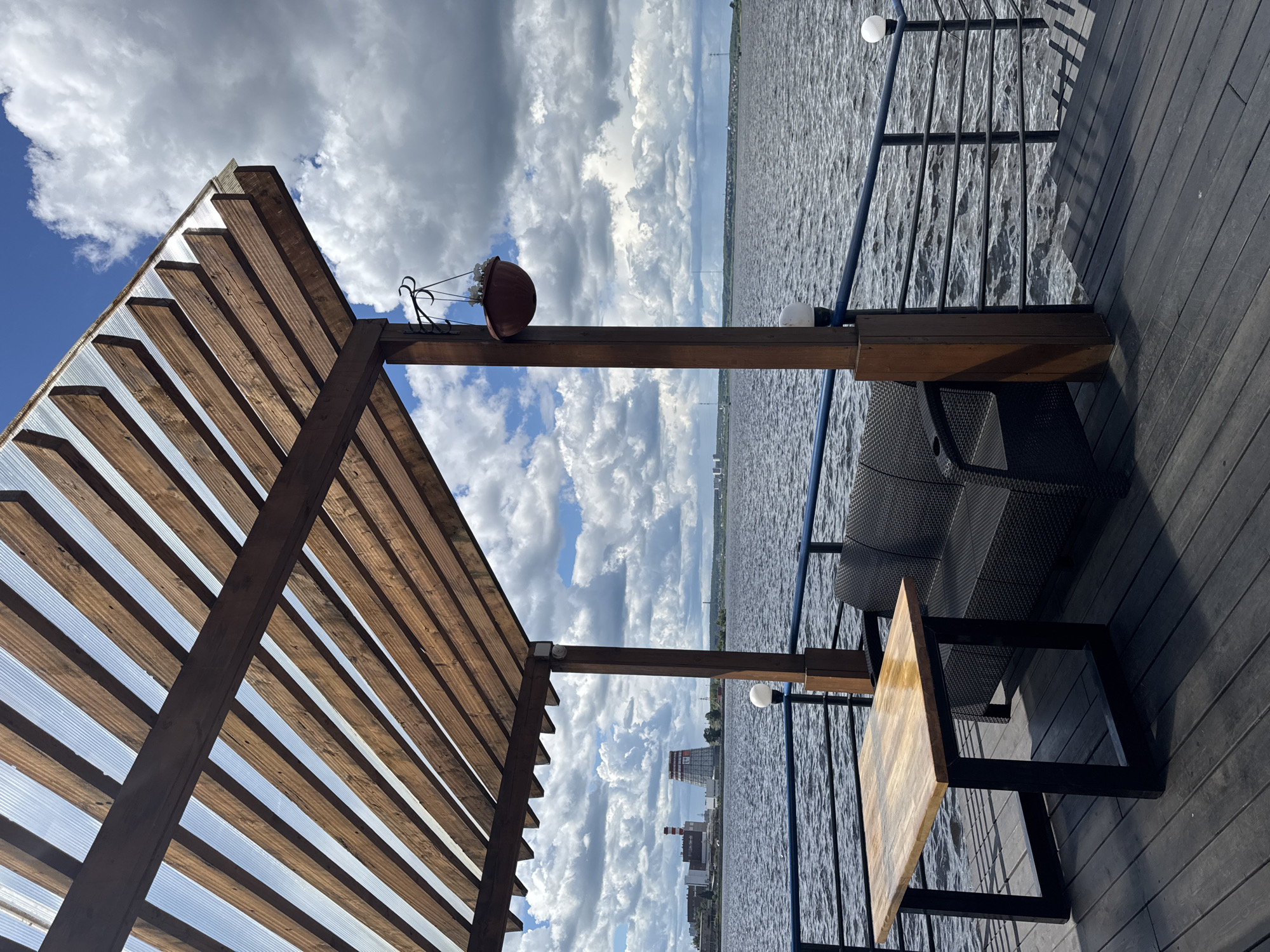 The image shows a wooden deck overlooking the water. A wooden pergola-like structure is visible on the left side of the image, and a table and chair are on the right. A metal railing separates the deck from the water. In the distance, buildings can be seen across the water. The image appears to be taken outdoors on a partly cloudy day. Given the location identified by the user, the location is likely the city of Izhevsk, Russia, potentially along the Izhevsk Pond or a similar body of water.