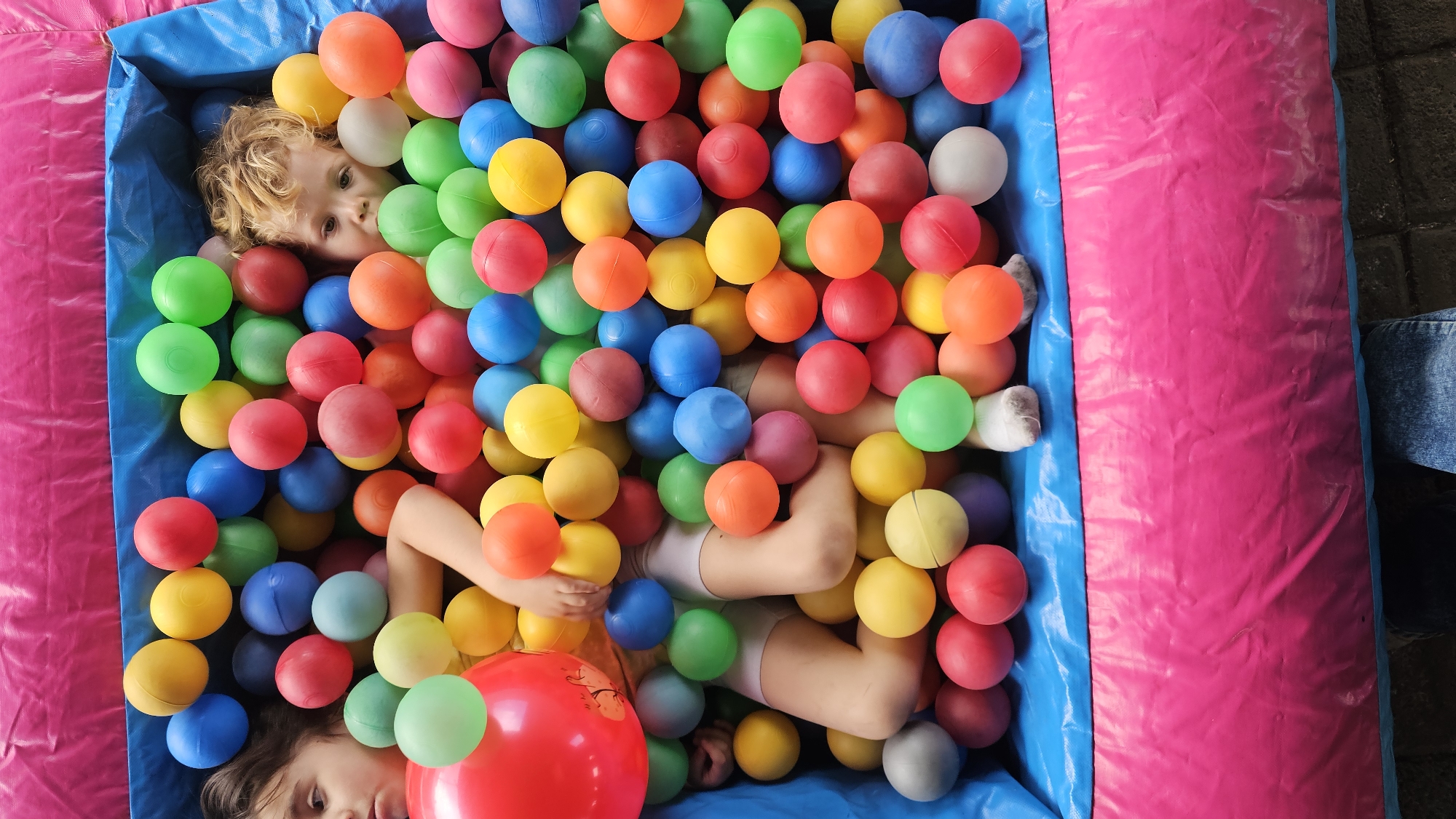 Two children are lying in a ball pit. The pit is lined with blue and pink material, and filled with various colored plastic balls including red, green, yellow, orange, blue, and white. One child with blonde hair is visible in the upper left, and another with brown hair is in the lower left. The brown haired child is partially covered by a large red ball. A person in denim is visible in the upper right. The setting is San Bernardino Tlaxcalancingo, Mexico.