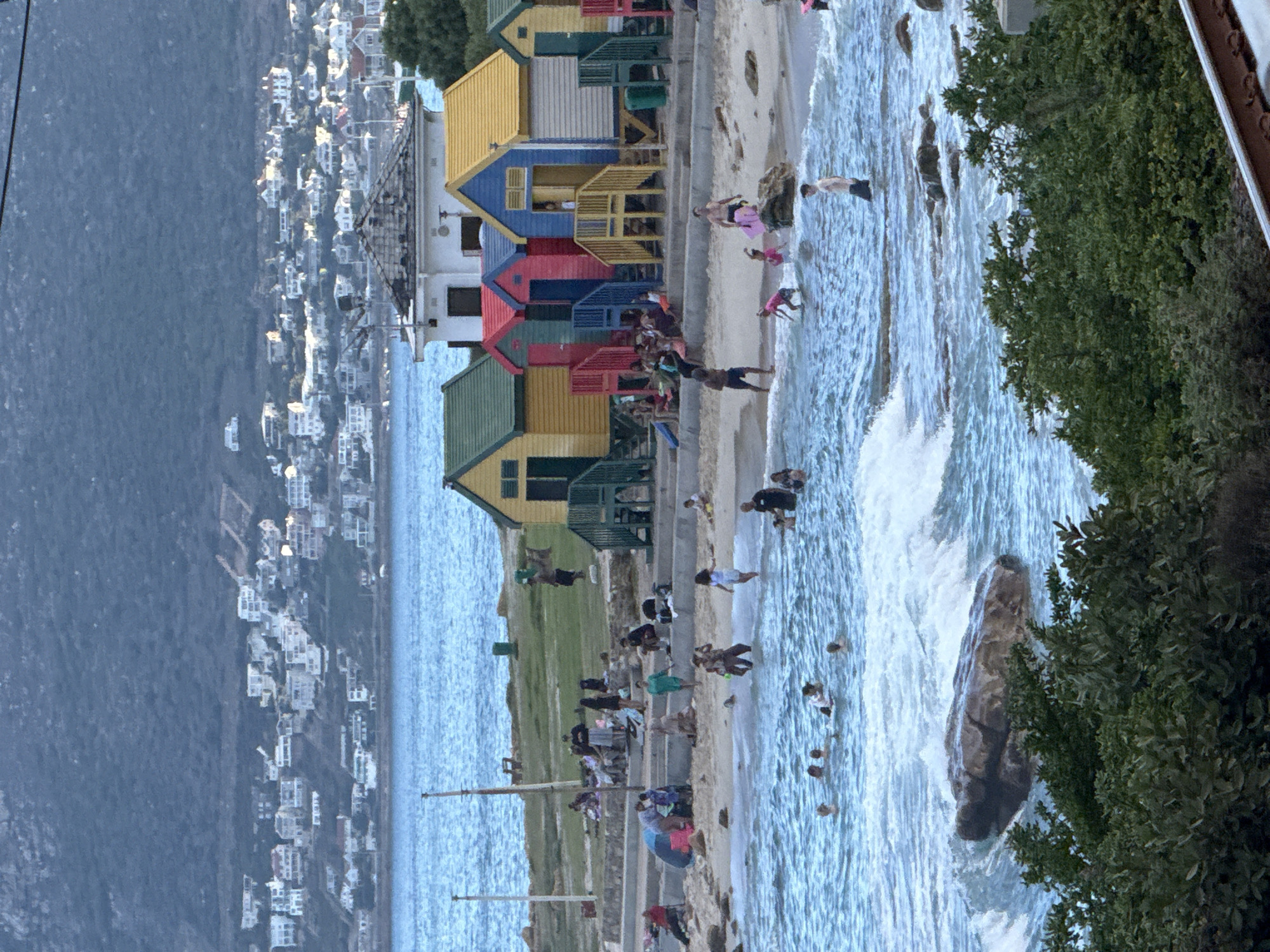 The image depicts a beach scene at Muizenberg Beach in Cape Town, South Africa. Several colorful, wooden beach huts line the shore, each painted in a different bright color. A group of people is spread across the beach and in the water. The water is churning with waves. On the far left, a cityscape is visible along the coast. Foliage frames the right side of the image.