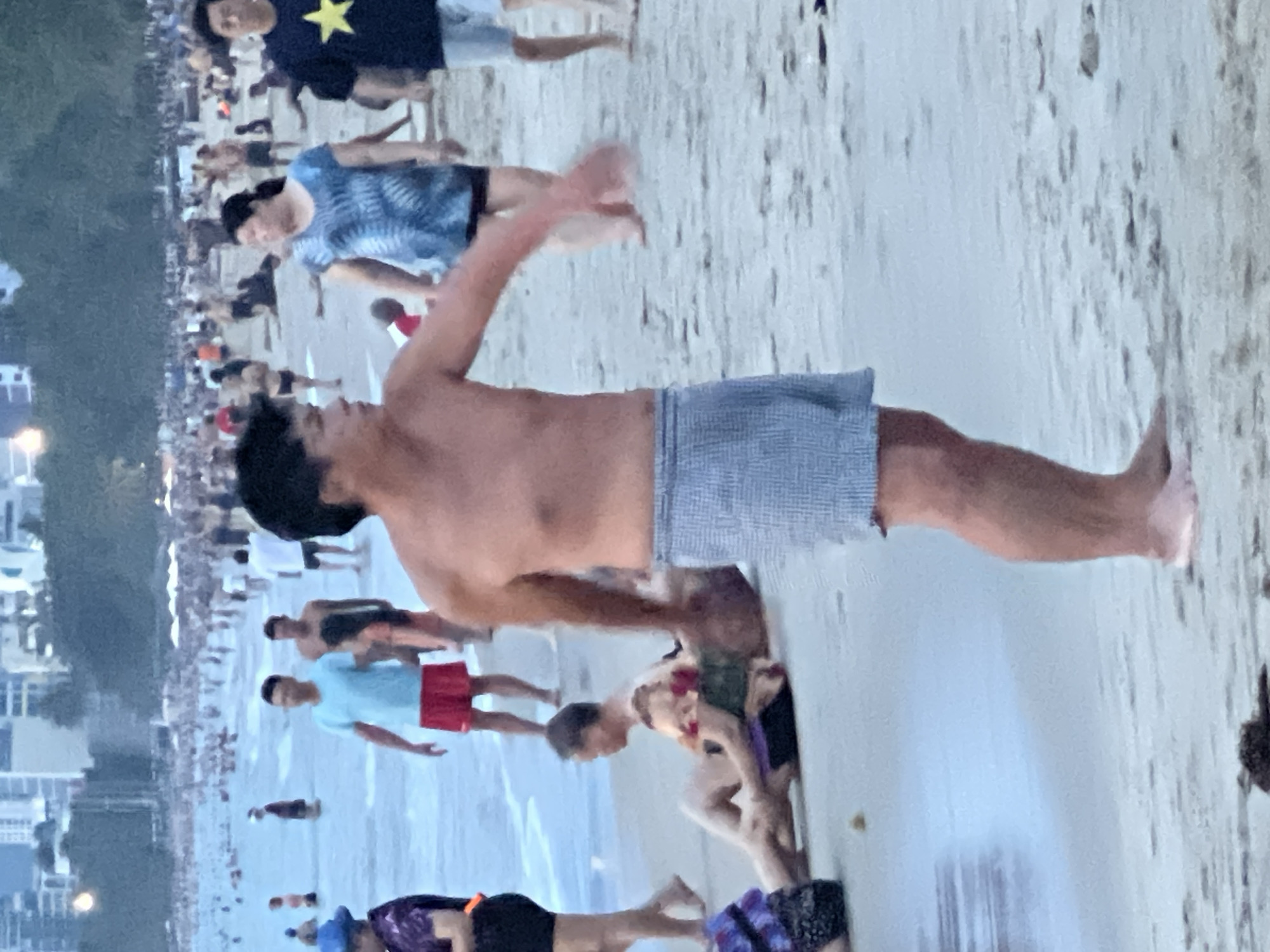 This is a medium-shot, eye-level, outdoor photograph of a crowded beach in Nha Trang, Vietnam. The foreground is dominated by a man standing shirtless, wearing blue and white checkered swim trunks. His body is positioned facing left, with his right arm extended outwards as if in mid-gesture. His facial features are not clearly discernible due to the image quality.
In the midground, a beach scene unfolds with numerous people scattered across the sand and in the shallow water. Several individuals are visible wading in the ocean, while others are relaxing or walking on the beach. A man in a light blue t-shirt and red shorts stands in the water. Further in the background, a woman in a blue and white patterned shirt is walking, and to her right, a man wearing a dark t-shirt with a yellow star emblem and light blue shorts is walking towards the right.
The background reveals the urban landscape of Nha Trang, with several tall buildings visible against a hazy sky, suggesting a coastal city environment. The overall atmosphere is one of a bustling public beach with many people enjoying themselves. The water appears calm and slightly choppy at the edge. The sand is light brown and shows signs of being wet in the foreground. The lighting suggests it could be late afternoon or early evening, with a diffused light.