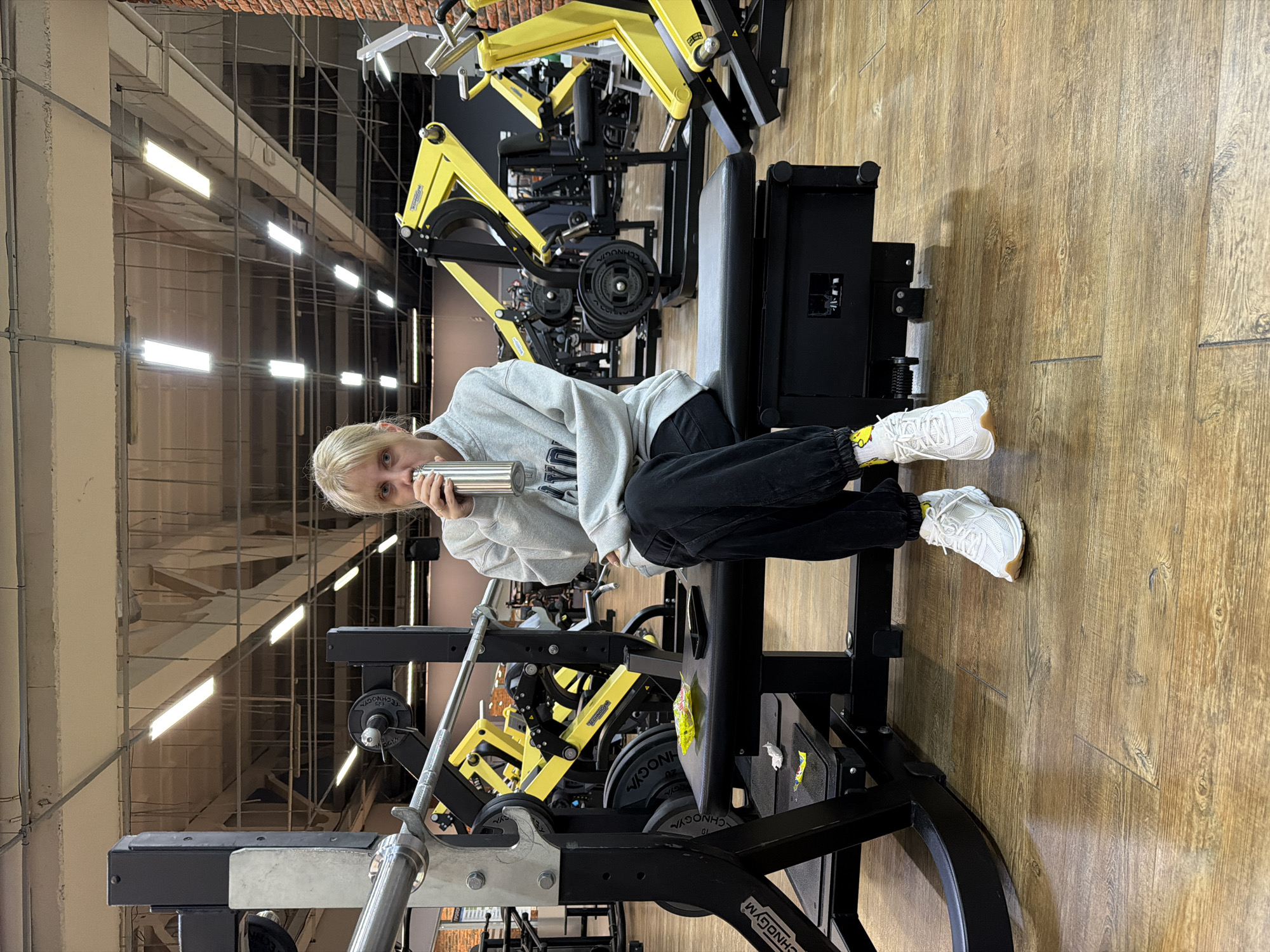 A person with short blond hair is seated on a piece of gym equipment in a gym. The person is holding a silver-colored cylindrical object near their mouth. The gym has multiple exercise machines with yellow and black frames and weights. The floor is made of wood. The gym is brightly lit by rows of fluorescent lights hanging from the ceiling. The city is Chaikovsky, Russia.
