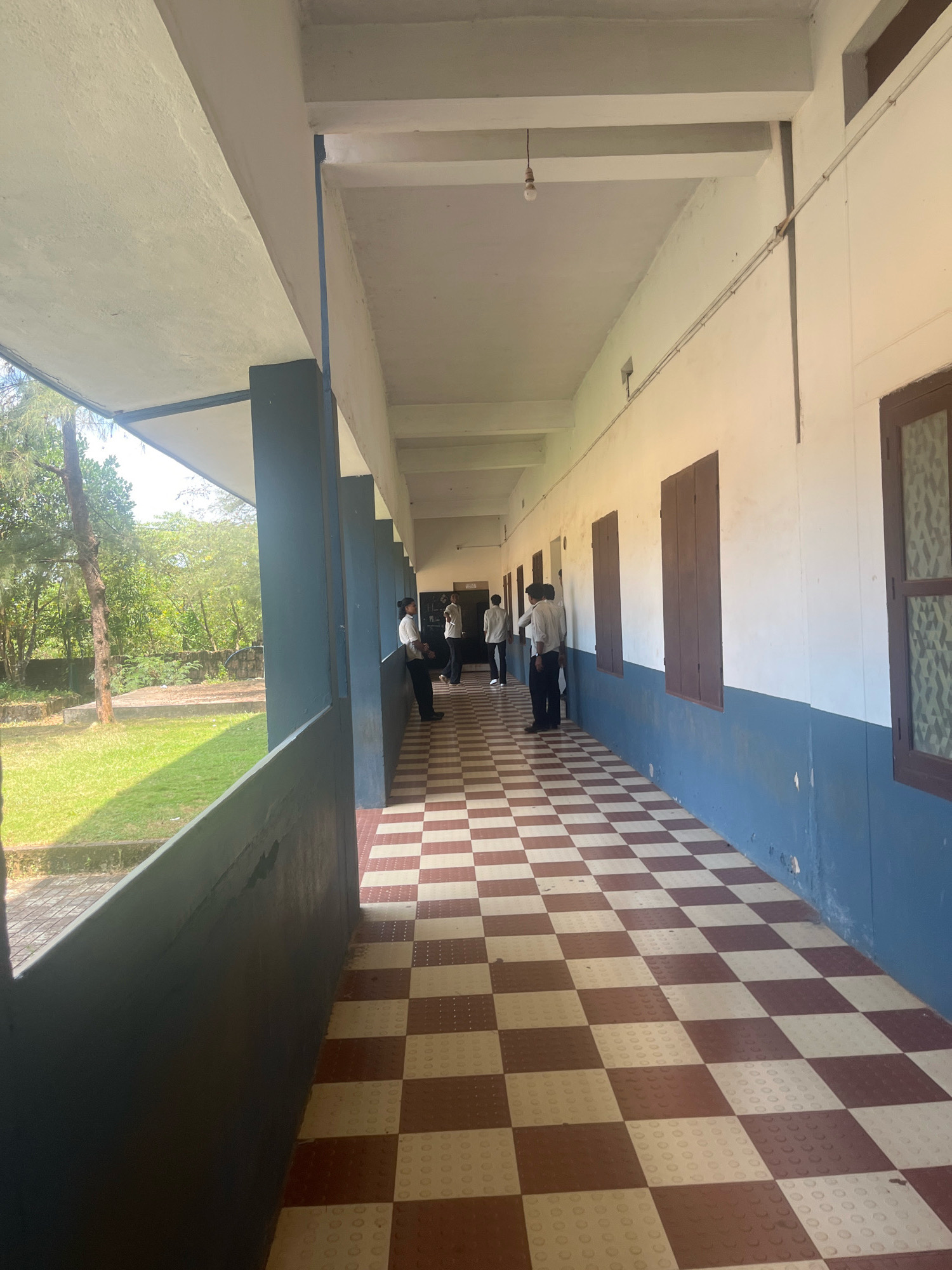 The image depicts a covered outdoor walkway or verandah of a building in Malappuram, India. The walkway has a checkered floor with alternating red and white tiles. The left side of the walkway is bordered by a dark grey wall, beyond which is a grassy area with trees and some vegetation, suggesting an outdoor or garden setting. The right side of the walkway is an interior wall of a building, painted white and blue. This wall features several dark brown wooden-framed windows with shutters. Several individuals, appearing to be young men in white shirts and dark trousers, are standing or walking along the walkway, some interacting with each other. An exposed light bulb hangs from the ceiling of the walkway in the distance. The architecture suggests a school, college, or other institutional building.