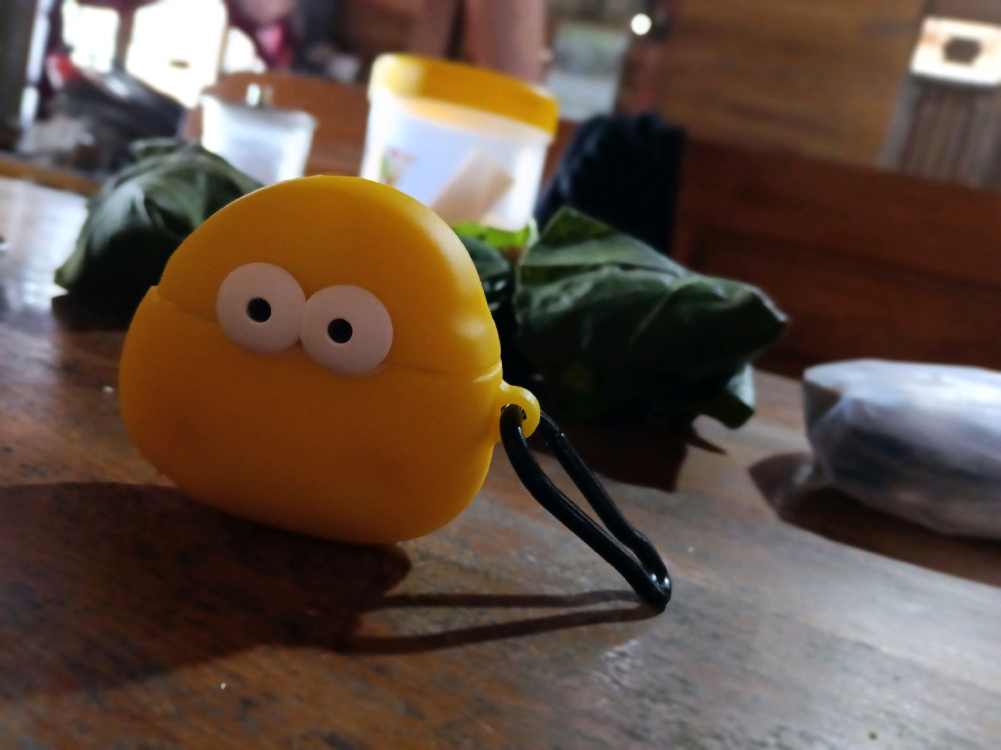 A yellow, cartoonish object with two large white eyes and black pupils rests on a wooden table. It has a black loop attached to its side. In the background, out of focus, are various objects including a white plastic container with a yellow lid, and some green leafy items. The scene appears to be indoors, possibly in a residence. The image was taken in Galuk, Indonesia.