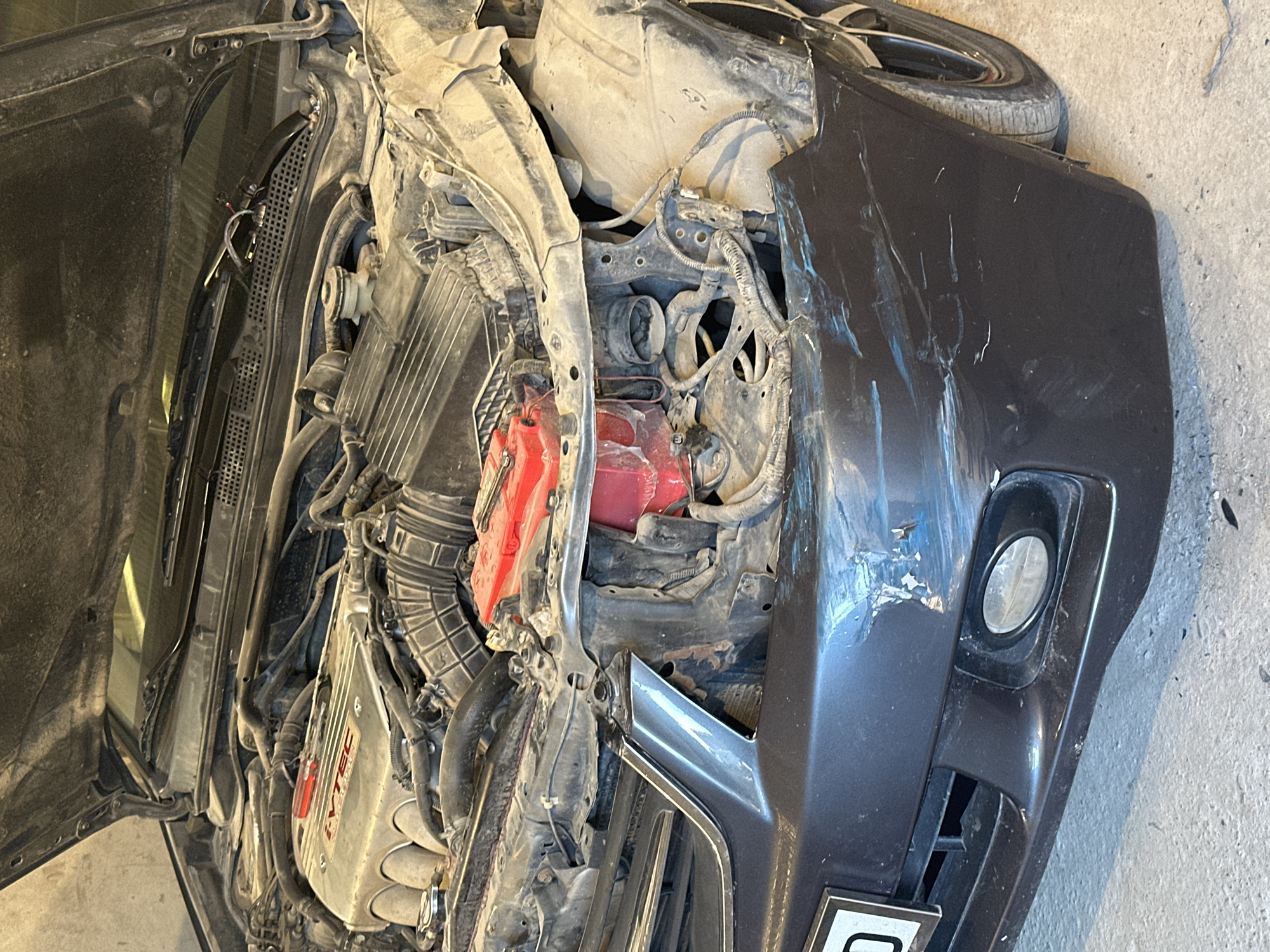 The image shows the front end of a dark-colored sedan, heavily damaged. The hood is open, revealing the engine compartment, which is dusty and cluttered with wires and hoses. The front bumper is scuffed and dented, with visible paint damage. The headlight on the right side is missing, and the surrounding bodywork is deformed. A red battery is visible in the engine bay. The car appears to be stationary in what looks like a garage or workshop, with a concrete floor and a light-colored wall in the background. The location is International, Kyrgyzstan.