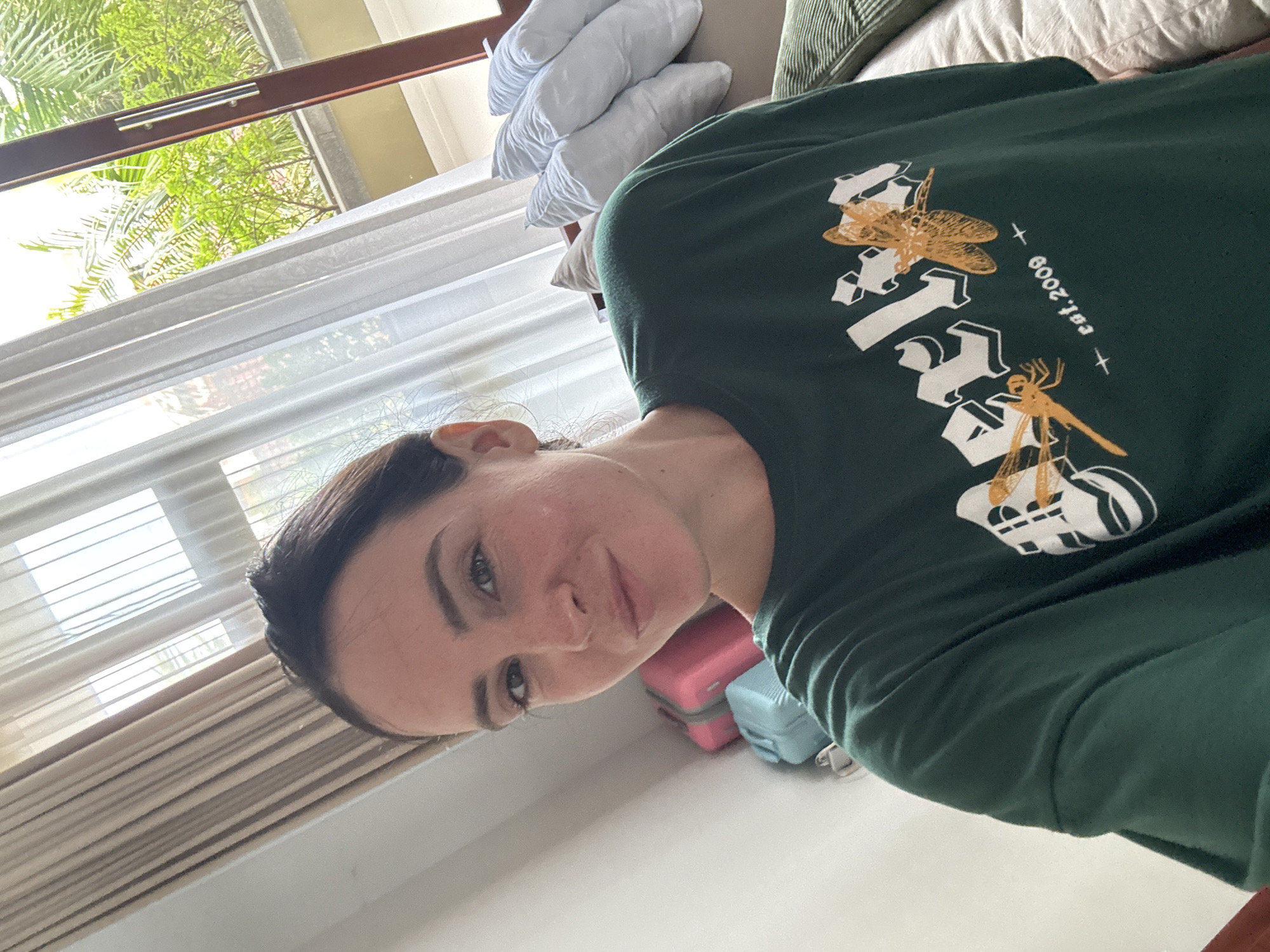 The image is a selfie taken indoors, likely in a hotel room or apartment in North Kuta, Indonesia, as suggested by the tropical foliage visible through the window. The subject, a woman with brown hair pulled back, is wearing a dark green t-shirt with a graphic design featuring the word "Merlin" and dragonflies. She is looking directly at the camera with a slight smile. In the background, a window is partially covered by sheer white curtains and beige drapes, allowing natural light to illuminate the scene. To the left of the woman, a stack of suitcases, one pink and one light blue, is visible. To her right, a couch is partially seen with several light-colored pillows and a green corduroy object, possibly a cushion or blanket. The overall impression is a casual and relaxed setting.