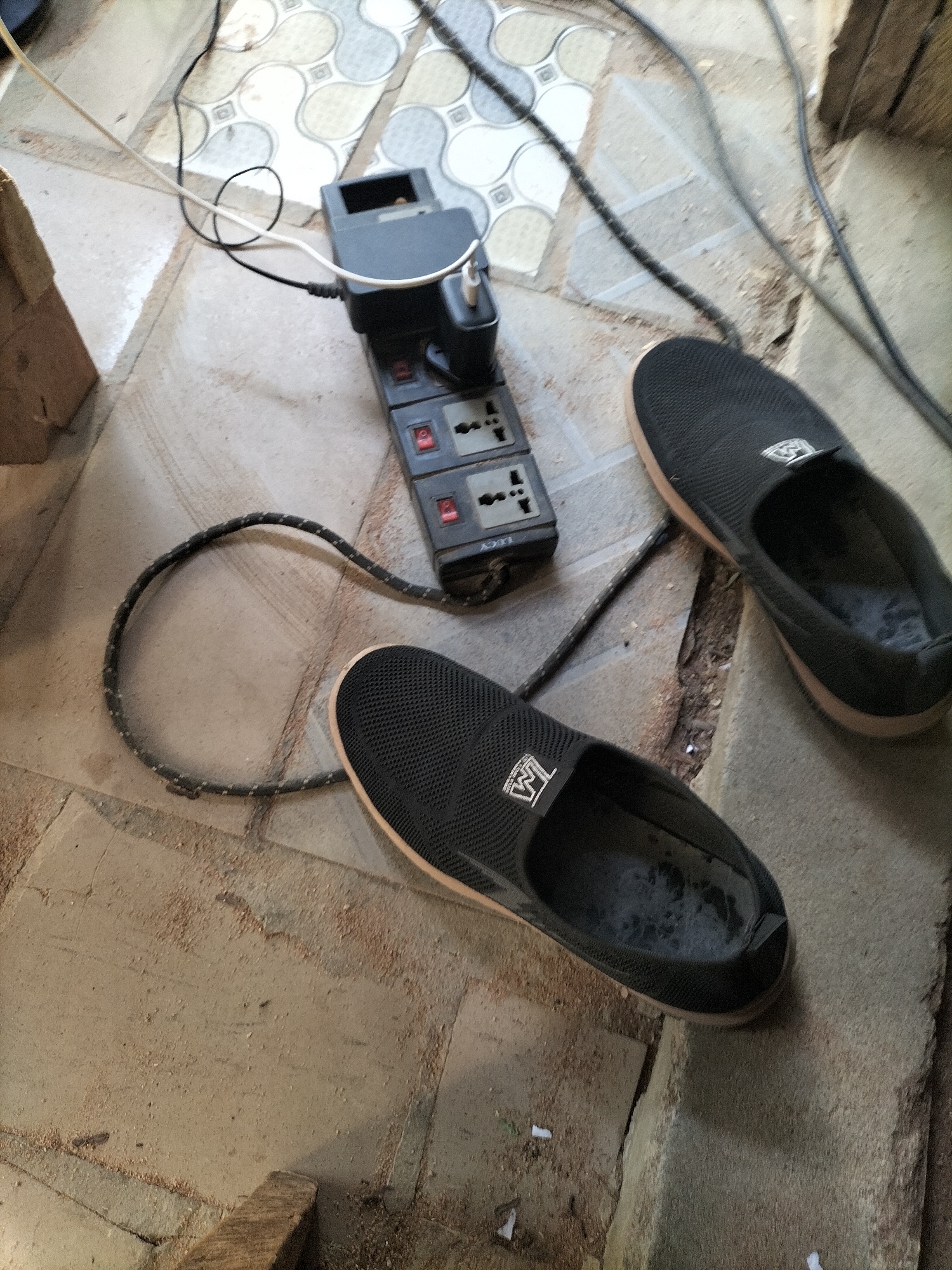 In Jalingo, Nigeria, on a floor tiled with a grey and white geometric pattern, an electrical power strip with multiple outlets and a black adapter is connected to a white power cord that loops near a black extension cord. Two black slip-on shoes are positioned on the floor. One shoe is to the right of the power strip, and the other is in the foreground, closer to the viewer. The floor is dusty and appears to be under construction or renovation, with scattered wood debris.