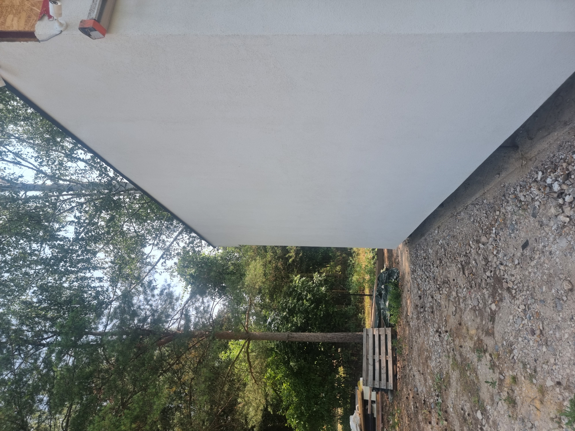 An exterior medium shot shows the side of a white, stucco-finished building with a dark soffit. The building is situated in a wooded area with tall trees and dense green foliage visible behind it. The ground in the foreground is rough and covered with gravel and some scattered pinecones. To the left of the building, wooden pallets and a dark tarp are stacked on the ground. The sky is overcast and partially obscured by tree branches. The scene appears to be in a rural setting, possibly in the city of Szczaki, Poland, indicating an outdoor construction or renovation site.