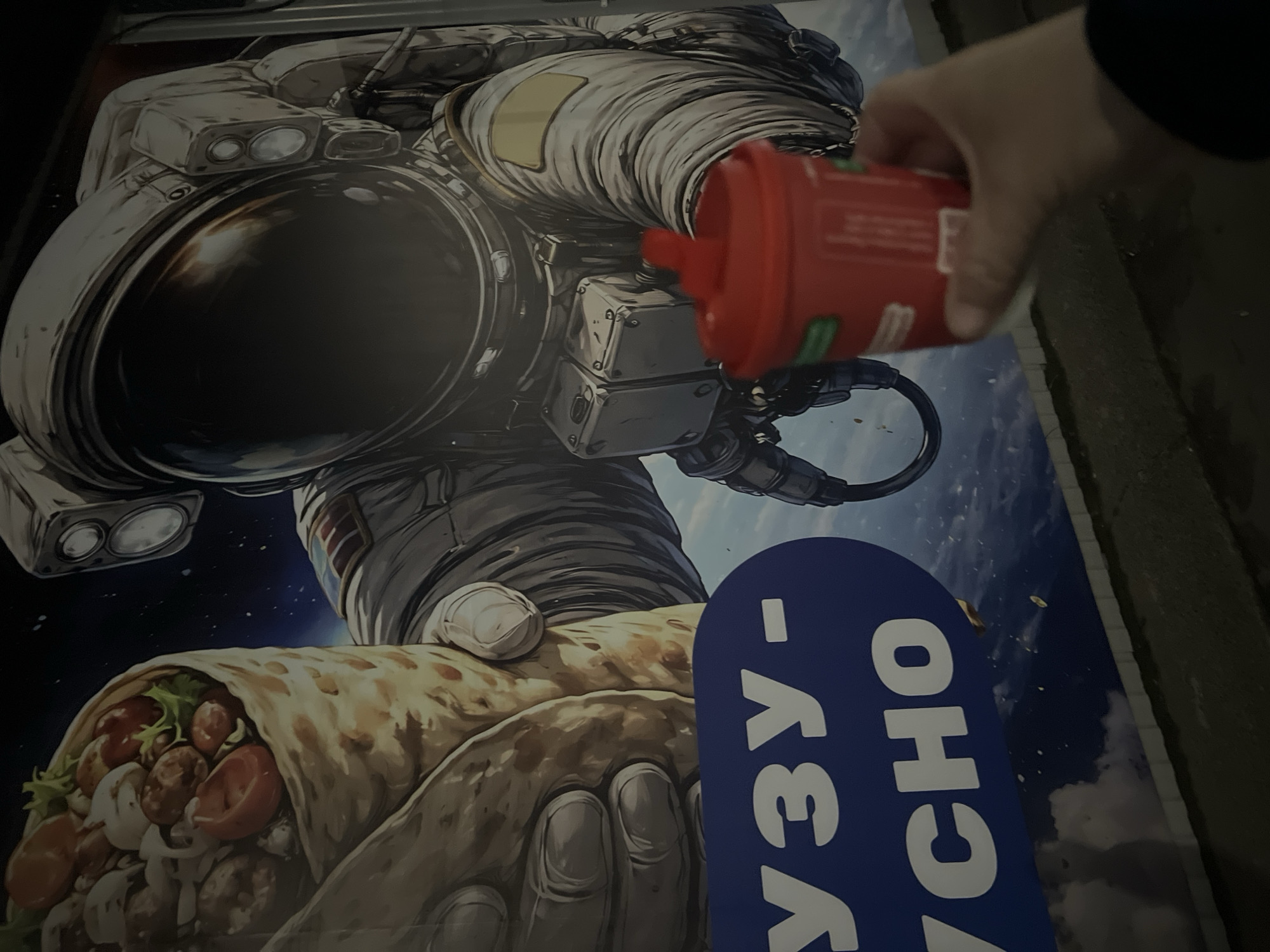 The image displays a nighttime outdoor scene in Chaikovsky, Russia, focused on a large advertisement poster featuring an astronaut. The poster depicts a realistically rendered astronaut in a white spacesuit with a dark, reflective visor, set against the backdrop of space with Earth or clouds visible below. The astronaut is prominently holding a large, well-filled wrap, resembling a burrito or shawarma, containing visible ingredients such as meatballs, mushrooms, tomatoes, and lettuce.
A human hand, entering the frame from the right, holds a red plastic squeeze bottle with white text and green accents. The bottle is positioned in front of the astronaut's suit, obscuring part of its arm and torso. The background around the poster is dark, indicating it is nighttime. Partially visible Cyrillic text on a blue section of the poster reads "УЗУ-ВКУСНО", with "ВКУСНО" meaning "delicious" in Russian, strongly suggesting the advertisement is for food, likely the wrap depicted. The scene captures an interaction with street advertising under low light conditions.