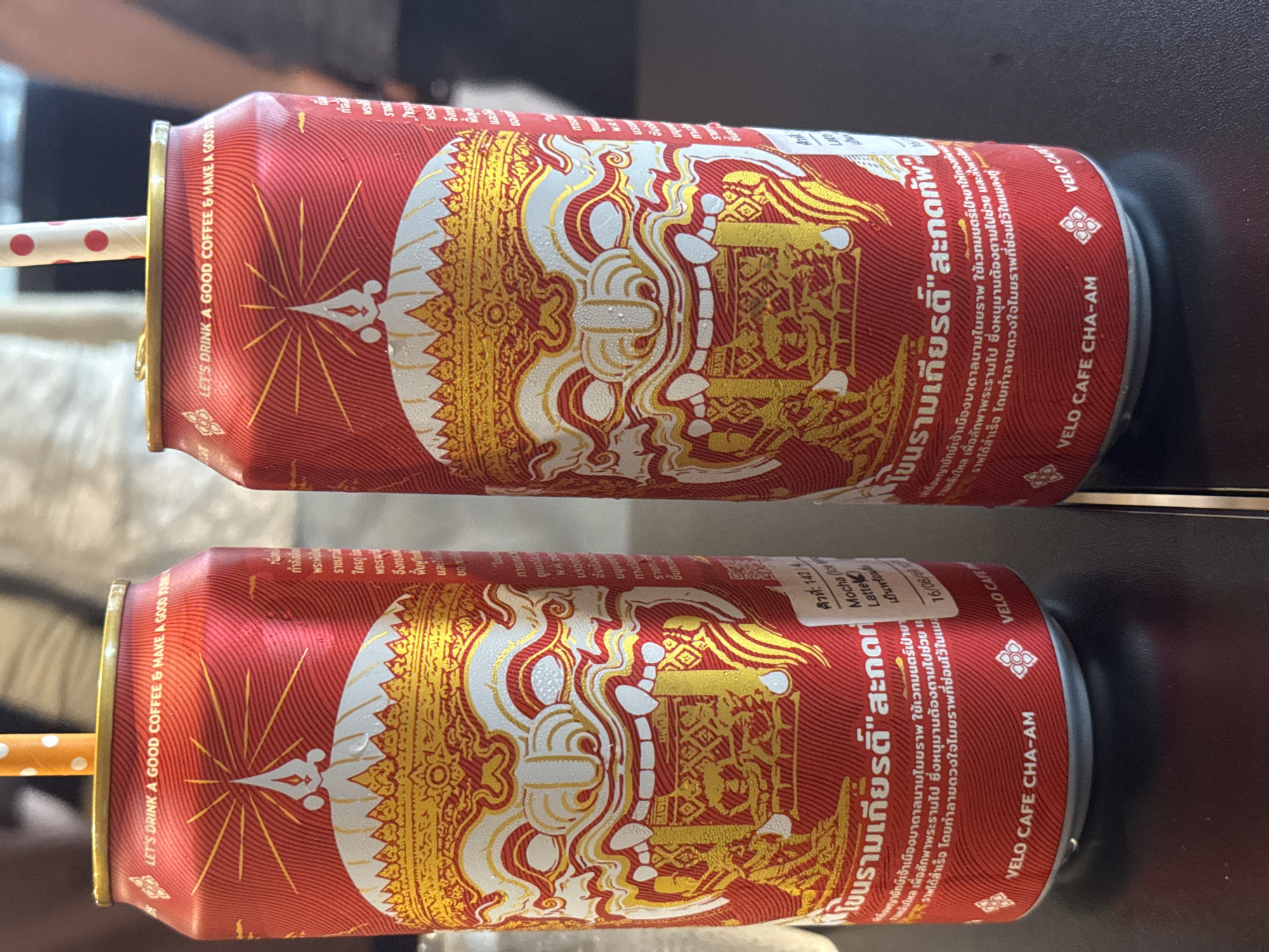 Two red cans of "VELO CAFE CHA-AM" coffee are displayed on a dark table, each with a straw. The cans feature a prominent design of a traditional Thai mask with intricate gold and white patterns. The text on the cans is in Thai and English. The text on the top of the cans reads "LET'S DRINK A GOOD COFFEE & MAKE A GOOD JOURNEY." The bottom of the cans has "VELO CAFE CHA-AM" printed on them. The image appears to be taken in an indoor setting, possibly a cafe in Hua Hin City Municipality, Thailand, given the branding. The lighting is natural and diffused, creating soft shadows.