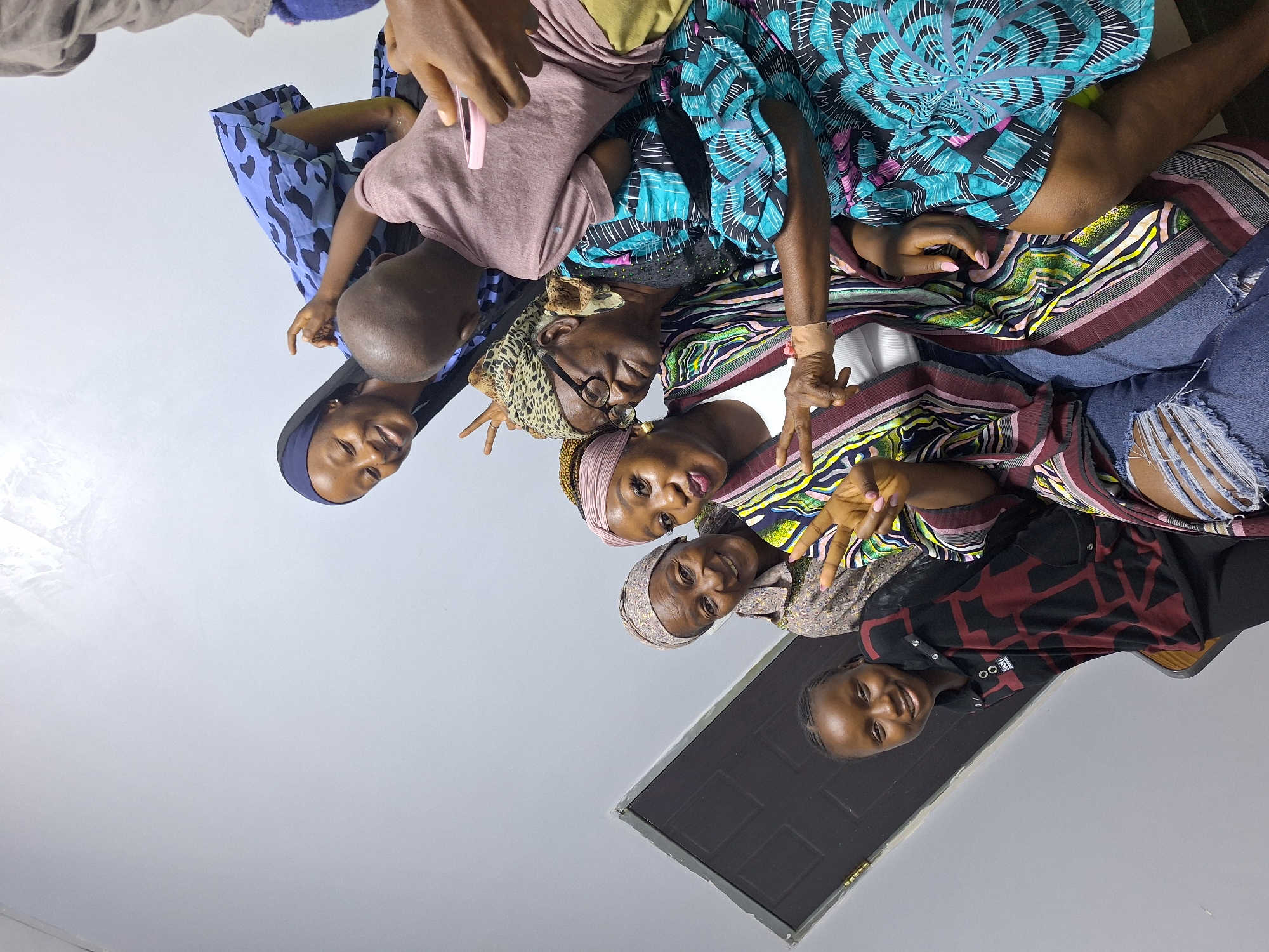 In Sabon Tasha, Nigeria, a group of six individuals, consisting of at least three generations of women and a young child, are gathered for a photograph. The scene appears to be indoors, possibly a home, with a plain gray wall dominating the background. A dark-colored door with a visible handle is on the left side of the frame.
The individuals are arranged in a cluster, with the youngest, a girl with braided hair, positioned on the lower left. She is wearing a black and red patterned long-sleeved shirt. To her right, a woman with a pink and white headscarf is making a peace sign with her fingers, her mouth slightly open. Next to her is an older woman wearing a patterned headscarf and glasses, looking towards the camera with a slight smile. She is adorned with a colorful, patterned garment.
Behind them, a young woman in a blue headscarf is leaning in, smiling and also making a peace sign. She is wearing a blue and black patterned dress. To her right, a child with a bald head is reaching out, possibly interacting with someone outside the frame or leaning on another person. Another individual's arm is visible on the right, holding a pink mobile phone. The overall atmosphere suggests a moment of shared joy and connection among family members.