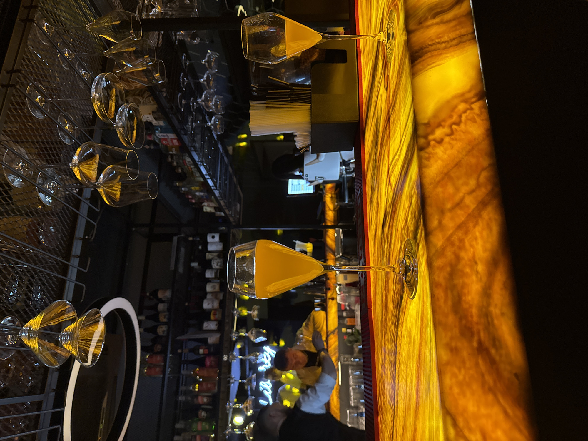 The image shows the interior of a bar or restaurant. A waiter is visible near a bar counter, which is illuminated with an amber-colored light. Two champagne glasses are filled with orange liquid, presumably a cocktail, and are placed on the bar top. Behind the bar, shelves are stocked with numerous bottles of alcohol. Wine glasses are neatly arranged in a rack overhead. In the background, various bottles and decorations can be seen, including neon signage. The location is identified as Perm, Russia.