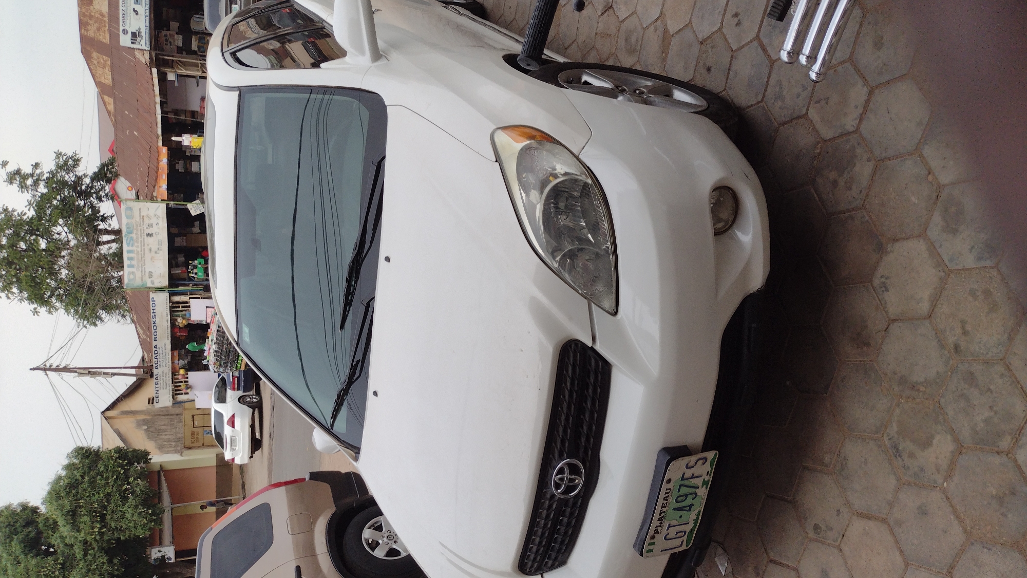 The image depicts the front of a white Toyota Matrix parked on a hexagonal paving stone surface. The vehicle has a Plateau State license plate, LGT-497FS. The scene includes a view of a street in the city of Jos, Nigeria, with commercial buildings visible in the background, including one that says "Central Agada Bookshop". Other vehicles are visible in the street. A tree is on the left.