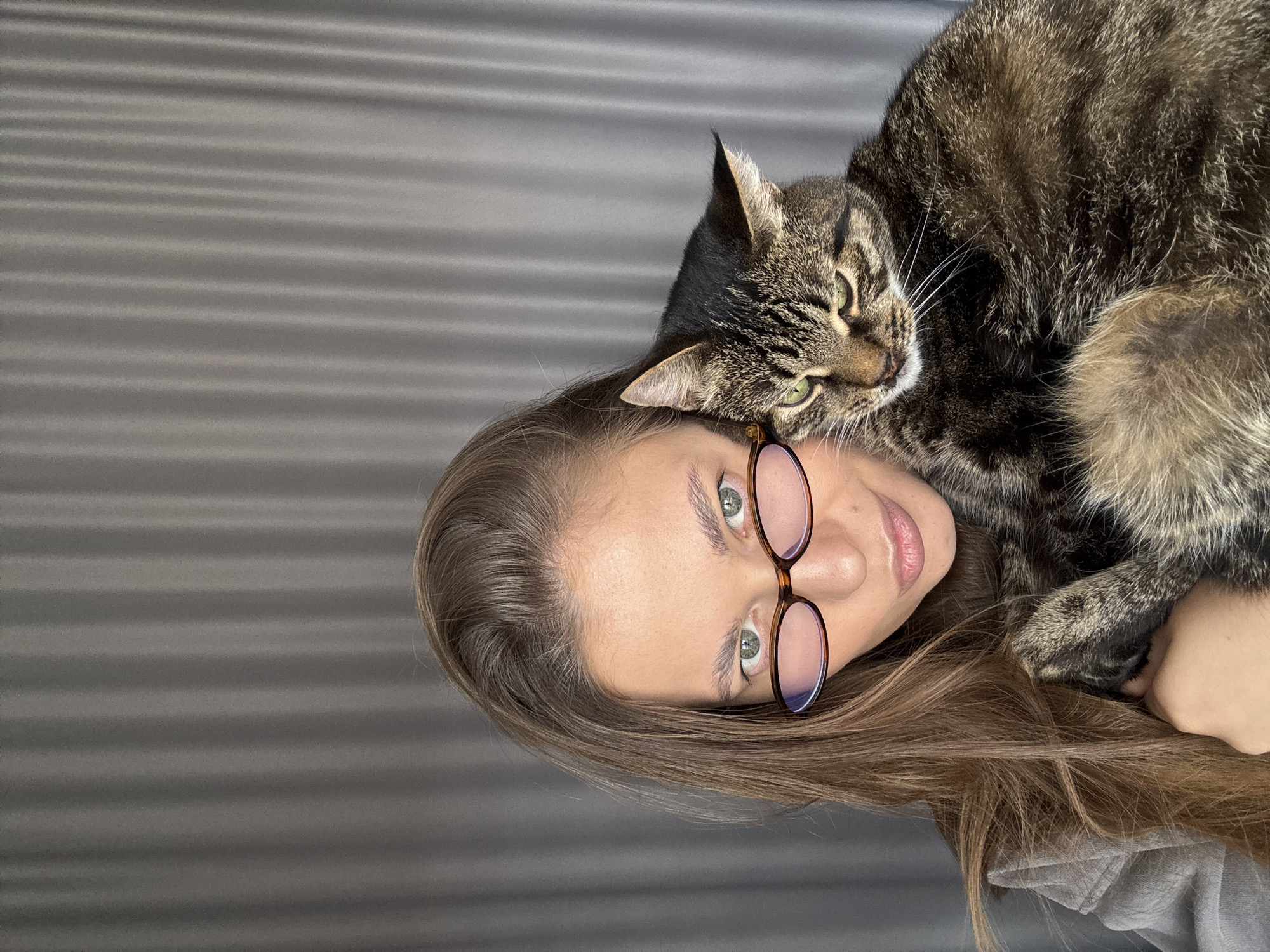 A vertically oriented photograph shows a woman with long brown hair and glasses with a brown frame lying on her back. A tabby cat rests on her head and face. The woman has fair skin and light-colored eyes. The background is a gray textured surface. The location is unknown, and there is no information indicating this photo was taken in Chernushka, Russia.