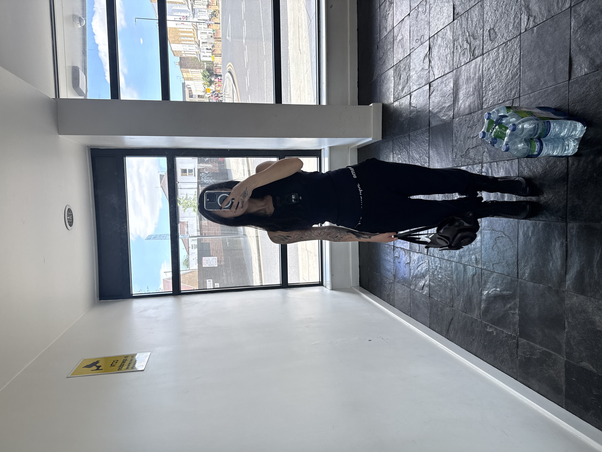 A person is standing in a brightly lit hallway with large windows that look out onto a city street. The hallway has white walls and a dark tiled floor. A yellow sign with black lettering, possibly a CCTV warning, is mounted on the left wall. The person is wearing black athletic wear, including a tank top and pants, and black boots. They are holding a black handbag and are taking a mirror selfie with their phone. A pack of six plastic water bottles is on the floor to the right of the person. Through the windows, a street scene with buildings, cars, and pedestrians is visible, consistent with an urban environment in London. The buildings appear to be terraced houses, typical of many London neighborhoods. The sky is blue with scattered clouds, suggesting a daytime scene.
