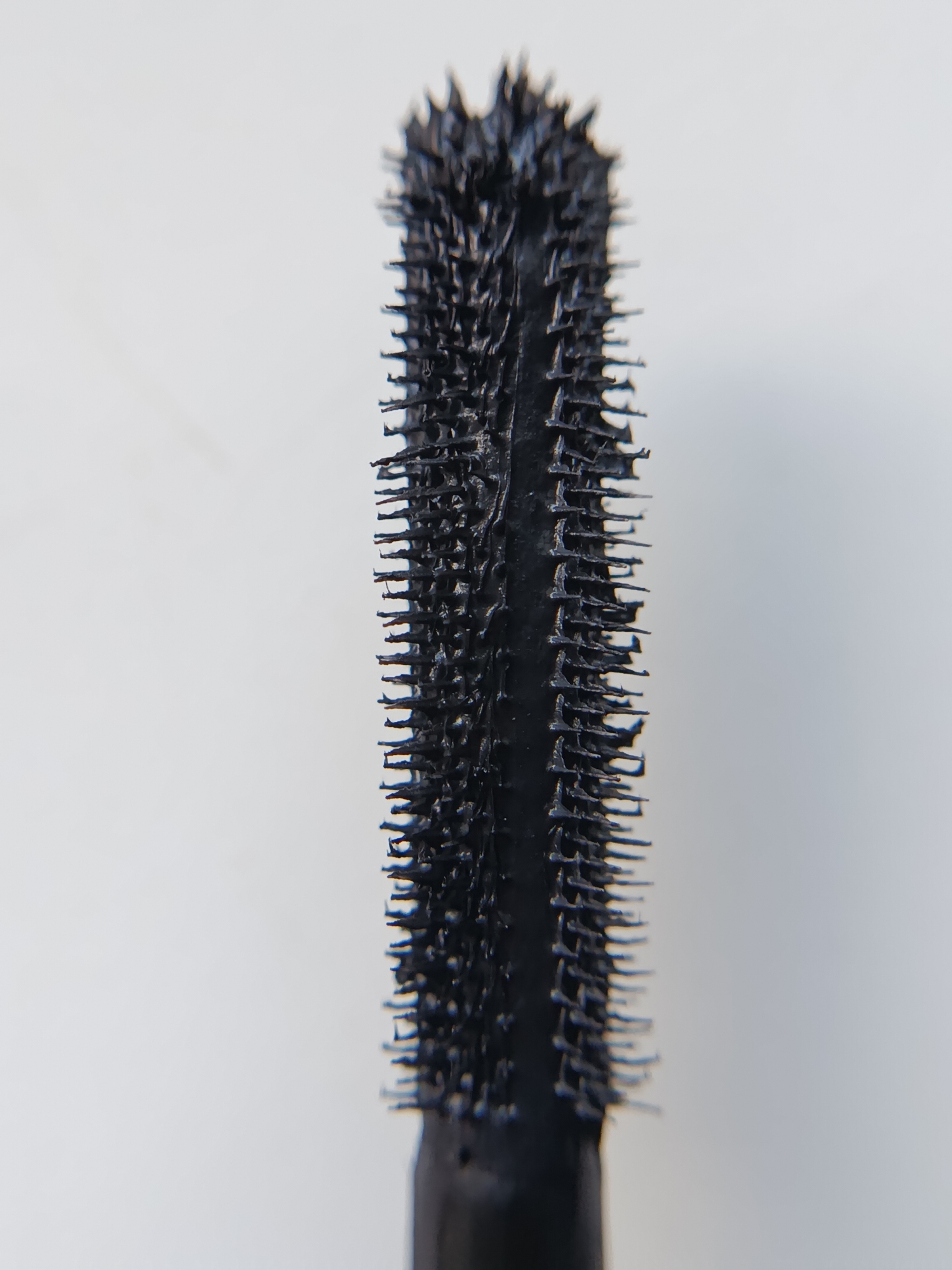 A close-up macro shot shows the black bristled wand of a mascara applicator against a plain white background. The bristles are dense and spiky, with a slight curve. The wand itself is black and appears to be made of plastic. The lighting is even, and the focus is sharp on the mascara wand, with the background slightly blurred.