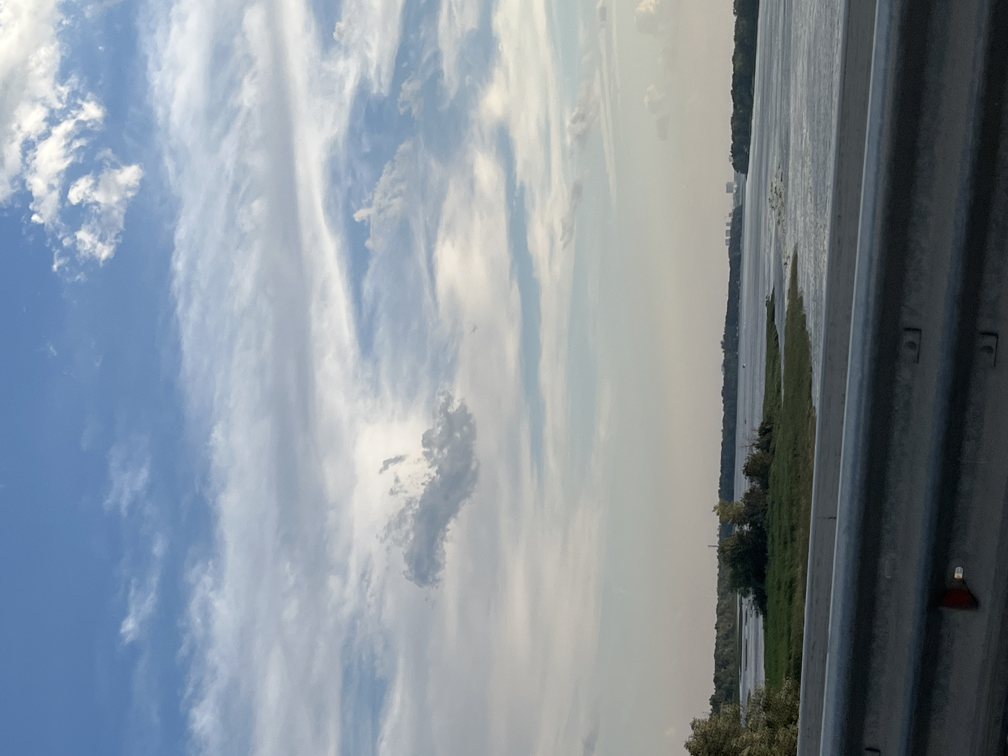 This is a vertical, slightly blurry photograph taken from a slightly elevated position, possibly a bridge or embankment, looking out over a body of water. In the foreground, a grey, corrugated metal barrier runs along the right side of the frame, suggesting the vantage point is alongside a road or pathway.
The dominant subject is the sky, which is a canvas of blue with a dynamic array of clouds. Wispy cirrus clouds streak across the upper half, interspersed with larger, puffier cumulus formations. One distinct, dark grey cloud in the center of the sky bears a peculiar, elongated shape, resembling a question mark or a stylized human figure. The lighting suggests it is daytime, likely late afternoon or early evening, as the sun appears to be casting a soft glow, illuminating the clouds from behind and creating a hazy, almost ethereal atmosphere.
The middle ground is occupied by a wide expanse of water, likely a river or lake, reflecting the soft light of the sky. The water's surface is calm, with gentle ripples. Along the far bank of the water, a line of trees forms a dense, dark green border, separating the water from the horizon. In the distance, on the horizon, a faint outline of buildings can be discerned, suggesting a settlement or town. The overall mood of the image is serene and contemplative, with the vastness of the sky and water evoking a sense of peace. The weather appears to be fair, with no immediate signs of rain. There is no visible text in the image.