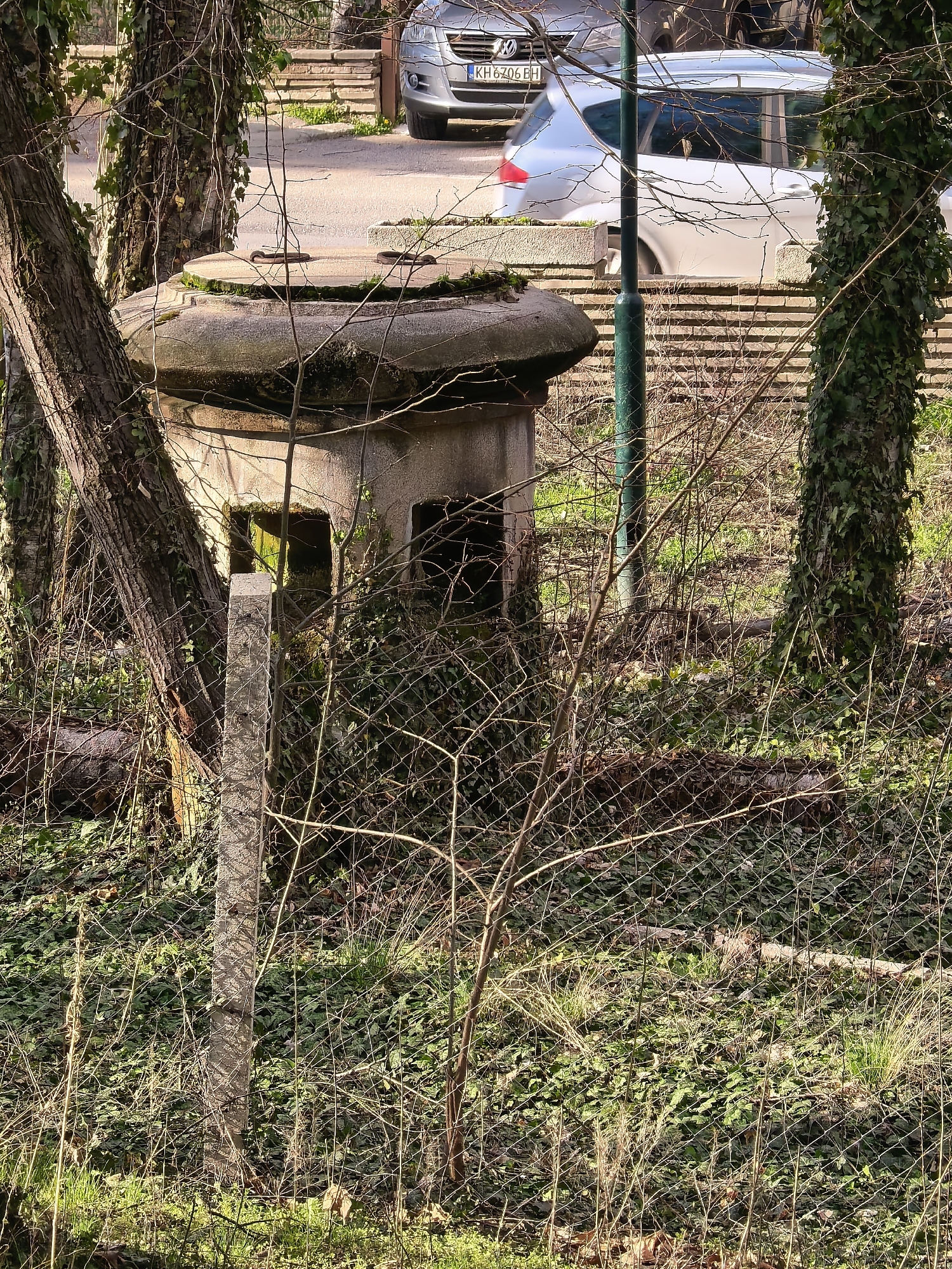 The image shows a concrete structure, possibly a bunker or an old pump house, situated among trees and overgrown vegetation behind a chain-link fence. The structure has a distinctive rounded top and small openings, possibly windows or entrances. A cement fence post is visible next to it. In the background, there's a street with parked cars, and one car has a license plate that reads "KH 6206 EH." The location appears to be an urban environment where nature has reclaimed a forgotten or abandoned structure. The city is unknown.