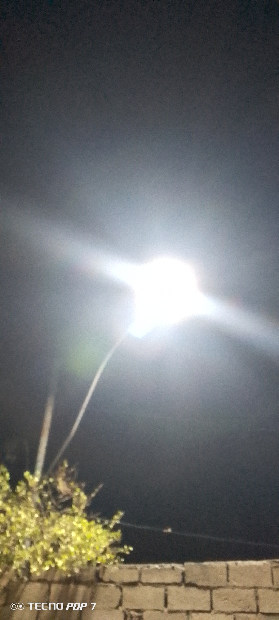 The image shows a night scene, likely in or near Altagracia, Venezuela. A bright streetlight dominates the center of the frame, causing significant glare and obscuring details around it. Below the light, there's foliage and the top of a brick wall. The sky above is dark. The words "TECNO POP 7" appear in the lower left corner, indicating the device used to capture the image.