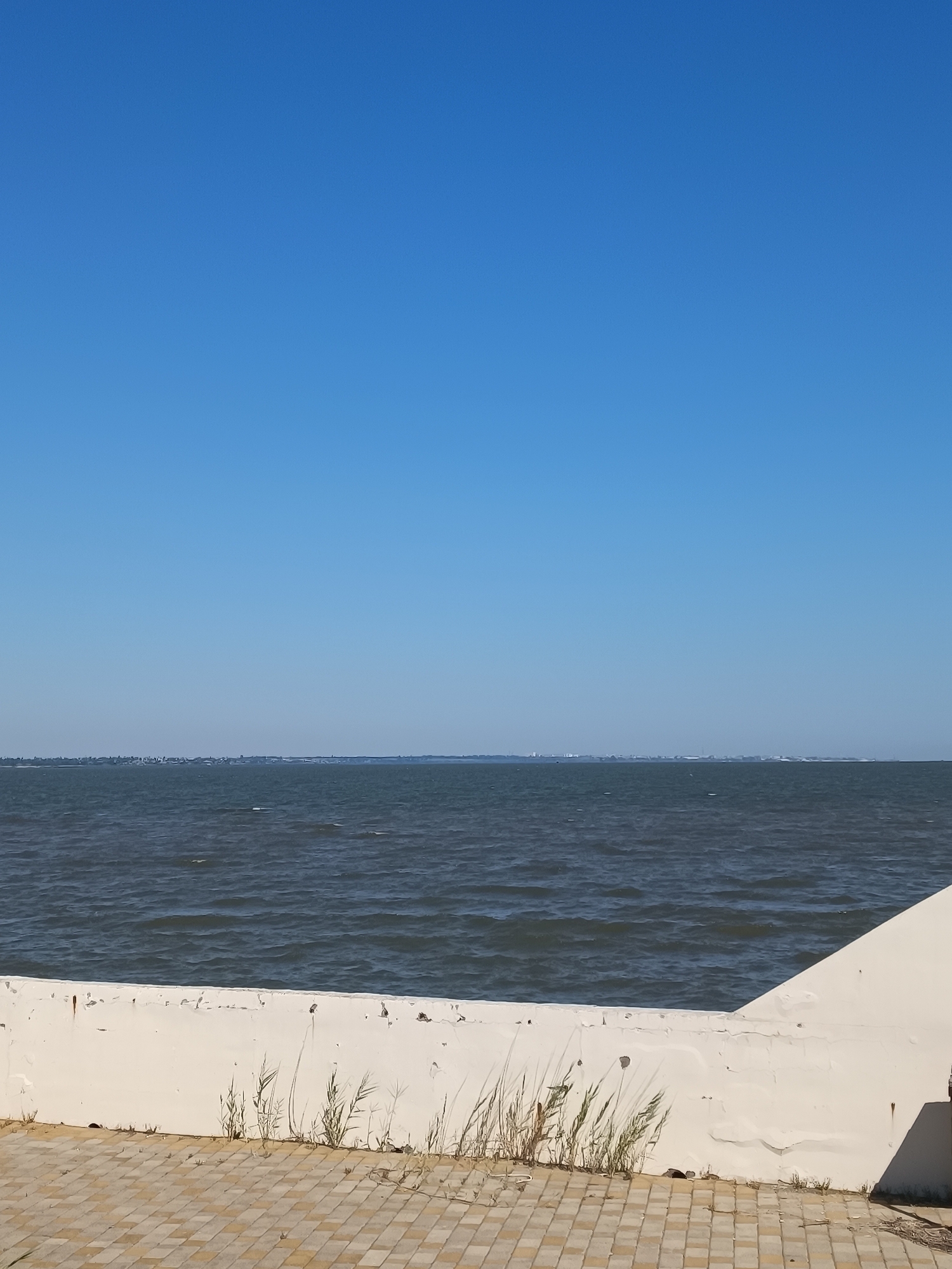 A serene, wide shot captures the vast expanse of the Black Sea under a clear blue sky. The horizon line, sharp and defined, separates the deep blue water from the light blue sky. A low, white concrete barrier runs horizontally across the foreground, showing signs of wear and tear with peeling paint and some rust marks. Small tufts of dry grass sprout from cracks and along the base of the barrier. To the right, the barrier meets a sloped, white wall that extends diagonally into the frame. The ground in front of the barrier is paved with light brown and beige square tiles laid in a diagonal pattern. In the distance, a faint, urban skyline is visible along the horizon. The overall impression is one of tranquility and a summer day by the sea.