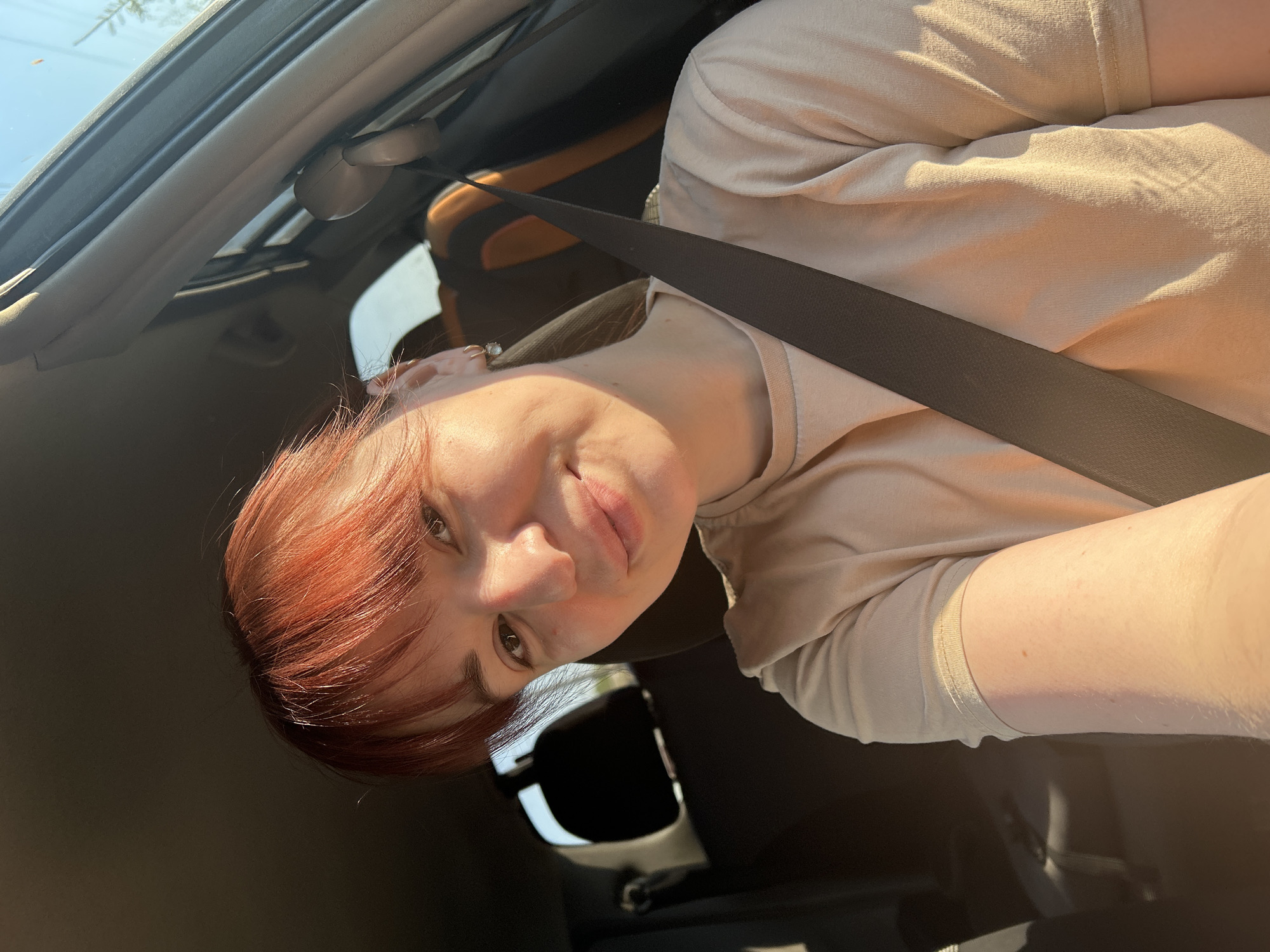 A woman with reddish-brown hair is wearing a seatbelt in a car with a sunroof. The photo is taken from a low angle, looking up at her face. She is wearing a light-colored shirt. No specific details are visible to suggest a location in Фоки, Russia.