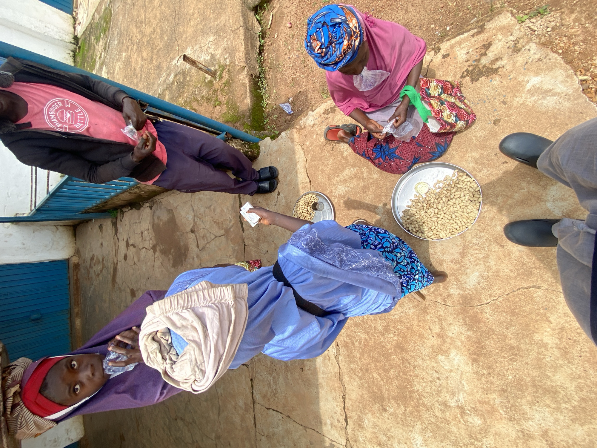 In Gura Top, Nigeria, four individuals are depicted outdoors on a concrete surface. One person, a young girl wearing a purple hijab and a red headscarf, is holding a clear plastic packet and appears to be drinking from it. Another individual, also in a hijab, is holding a packet in their hand and looking down. In the foreground, a person wearing dark boots and grey pants is partially visible. To the right, a woman in a pink garment and a blue headscarf is seated on the ground, holding a packet and a colorful bag. Several metal trays containing peanuts are scattered on the ground. The background features a building with blue doors and railings.