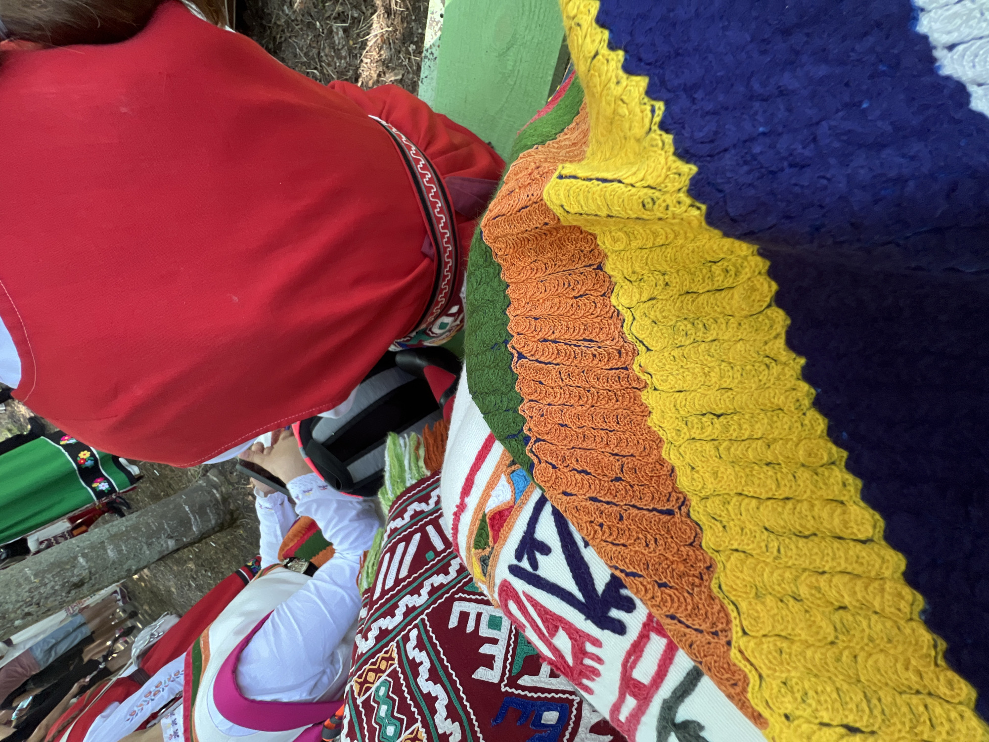 This image captures an outdoor scene in Bolyarovo, Bulgaria, centered on traditional textiles and individuals in customary attire. In the foreground, a stack of vibrant, hand-knitted or crocheted blankets is displayed, featuring distinct horizontal stripes in dark blue, yellow, orange, and green, each stripe exhibiting a textured, wavy pattern. Beneath these are other woven textiles, likely cushions or rugs, adorned with complex geometric patterns in white, red, green, and dark blue on maroon and white bases.
In the mid-ground, two individuals are prominent. On the left, a person is seen from the back, wearing a white long-sleeved shirt with embroidered cuffs, a white apron-like overgarment, and a colorful striped sash (pink, green, white). This individual holds and observes a dark-colored smartphone. To the right, a person is depicted from the upper back, wearing a bright red garment, possibly a vest or top, over a white layer, cinched by a narrow belt featuring white, black, and red geometric patterns. This person is positioned on a green painted wooden surface, possibly a bench or table.
The background contains several blurred figures, some standing, suggesting a gathering. Visible attire includes a green skirt with a decorative black band on a figure in the background right, and a red top with white embroidery on a figure in the far left background. A tree trunk is visible in the mid-ground, indicating a natural, outdoor setting with visible soil and scattered ground cover.