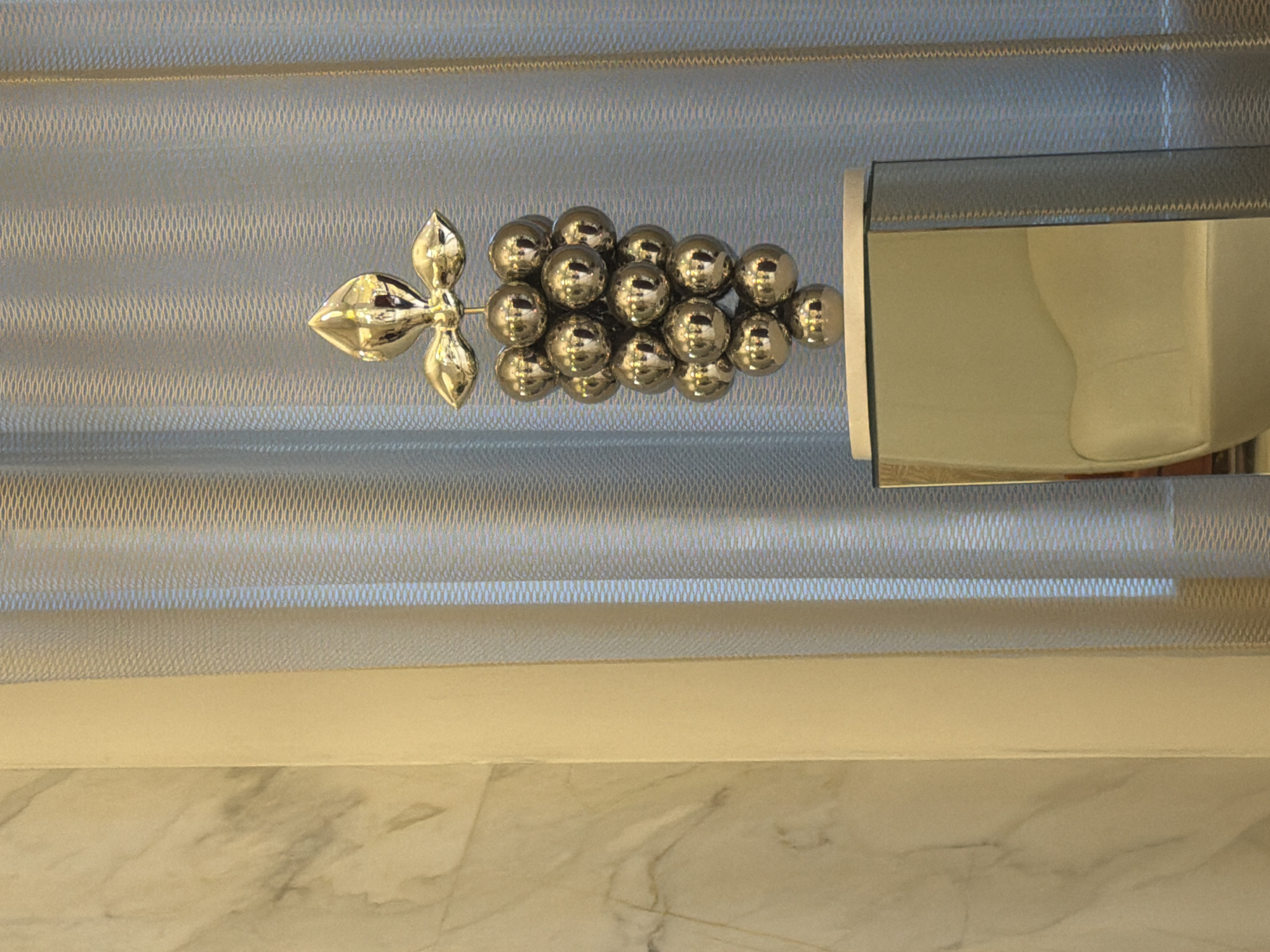 The image presents a close-up, vertically oriented view of an indoor setting, focusing on decorative and functional elements. In the center, a decorative item resembling a bunch of grapes is visible, crafted from numerous small, shiny, golden spheres. To the left of this cluster, a metallic, leaf-like or floral flourish extends, all appearing to be attached to a surface behind it.
This decorative element is set against a light-colored roller blind or window shade. The blind exhibits a subtle, intricate pattern, possibly a woven texture with fine horizontal lines and a criss-cross or diamond mesh design. To the right of the grape decoration, a reflective surface, potentially a mirror or polished metal fixture, is partially visible. This surface reflects a soft, light-colored, and curved form, possibly part of a larger fixture like a toilet or sink.
Below the blind, there is a horizontal band of solid, light beige or cream-colored material. The bottom section of the image reveals a polished, light-toned surface, appearing to be marble or a similar stone, characterized by subtle, natural veining. The overall lighting is soft and ambient, suggesting an indoor environment. No people or activities are present, and no text is visible. The scene implies a well-appointed room, possibly a bathroom, due to the combination of reflective surfaces and marble-like finishes.