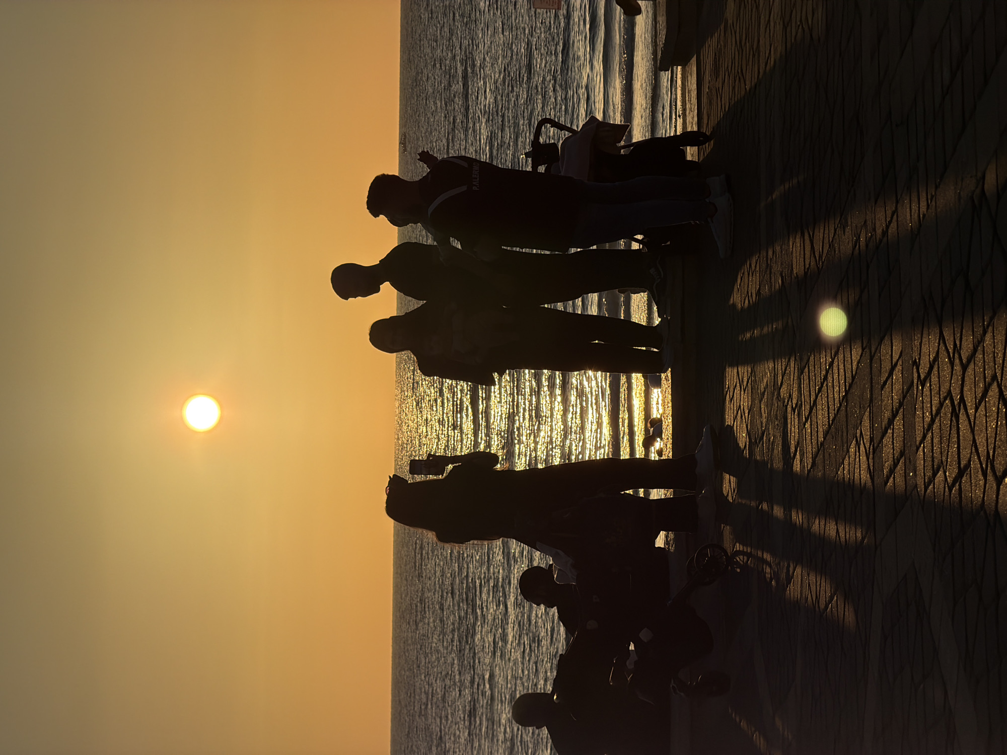 The image depicts a sunset scene on a seaside promenade in Tel-Aviv, Israel. Several people are captured in silhouette against the bright, golden-orange sky and the shimmering waters of the sea. The sun, a distinct white-yellow disc, is positioned low on the horizon, casting a warm, intense glow across the water, creating sparkling reflections.
Approximately eight to ten individuals are visible on the promenade. Most are standing, gazing towards the horizon, while a couple appear to be seated or crouching. Their figures are entirely silhouetted, obscuring facial features and most details, emphasizing their outlines against the vibrant backdrop. One individual, positioned towards the center-right, has the word "PALERMO" faintly visible in white text on the back of their dark shirt. Nearby, a stroller or small wheeled cart is also visible in silhouette.
The paved promenade extends along the right side of the frame, with long, dark shadows of the people stretching across its textured surface. A distinct, circular light flare or reflection is present on the ground in the lower right quadrant. The sky transitions from a bright golden hue near the sun to a softer orange further away. The overall atmosphere is peaceful and contemplative, indicative of a clear, calm evening by the sea.