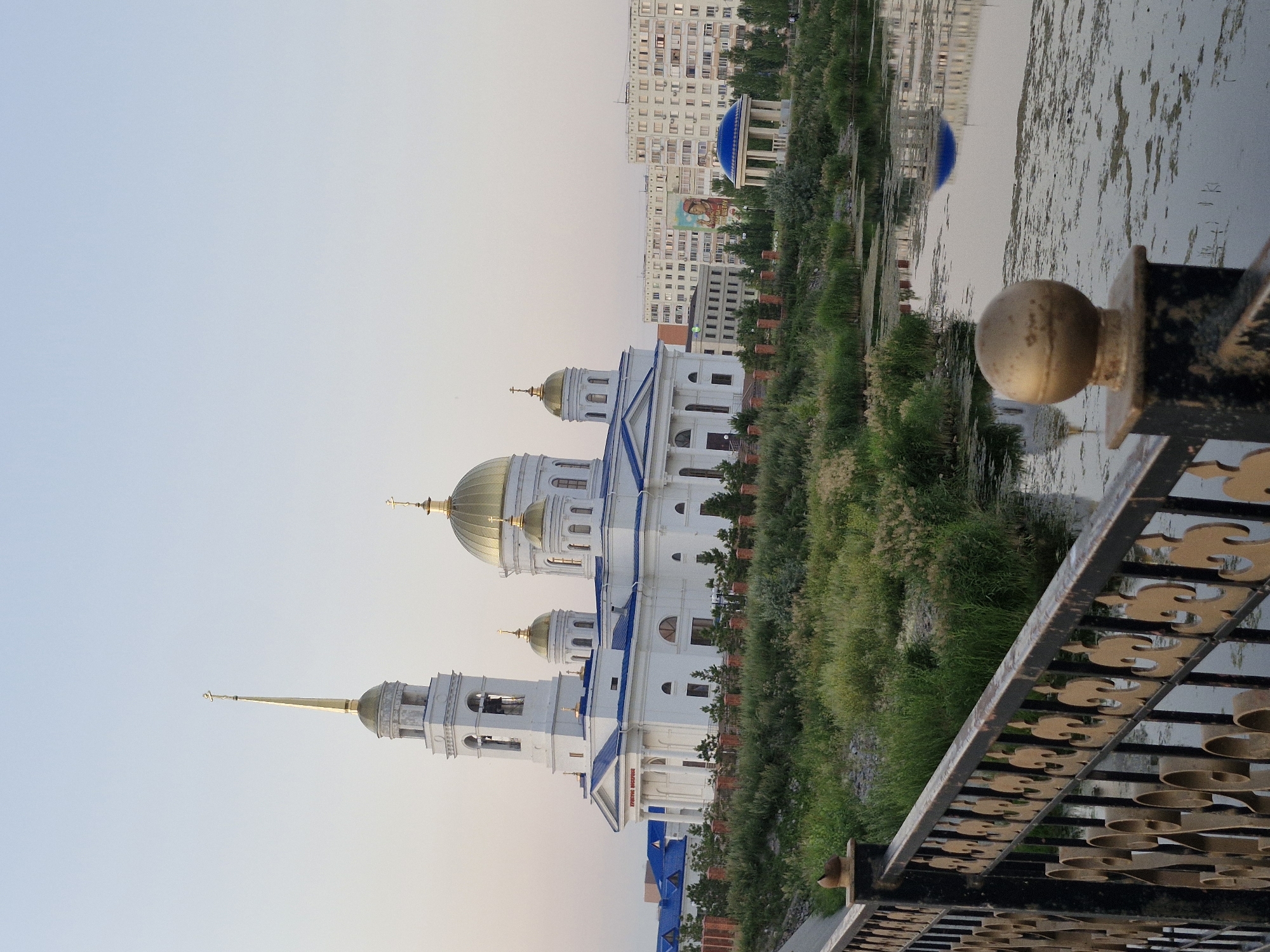 The image depicts a view of a white building with blue roofs and golden domes, likely a church or cathedral, situated near a body of water. Tall residential buildings and a blue-domed pavilion are visible in the background. The scene is bordered by a decorative black metal railing with golden scrollwork in the foreground, suggesting a public space like a park or promenade. The water reflects the sky and the surrounding structures, indicating a calm surface. Greenery, including trees and tall grass, lines the bank of the water. A billboard with a portrait is also visible on one of the buildings. The lighting suggests it is either early morning or late afternoon. There is no discernible activity or people in the frame. The overall mood is serene and architectural.