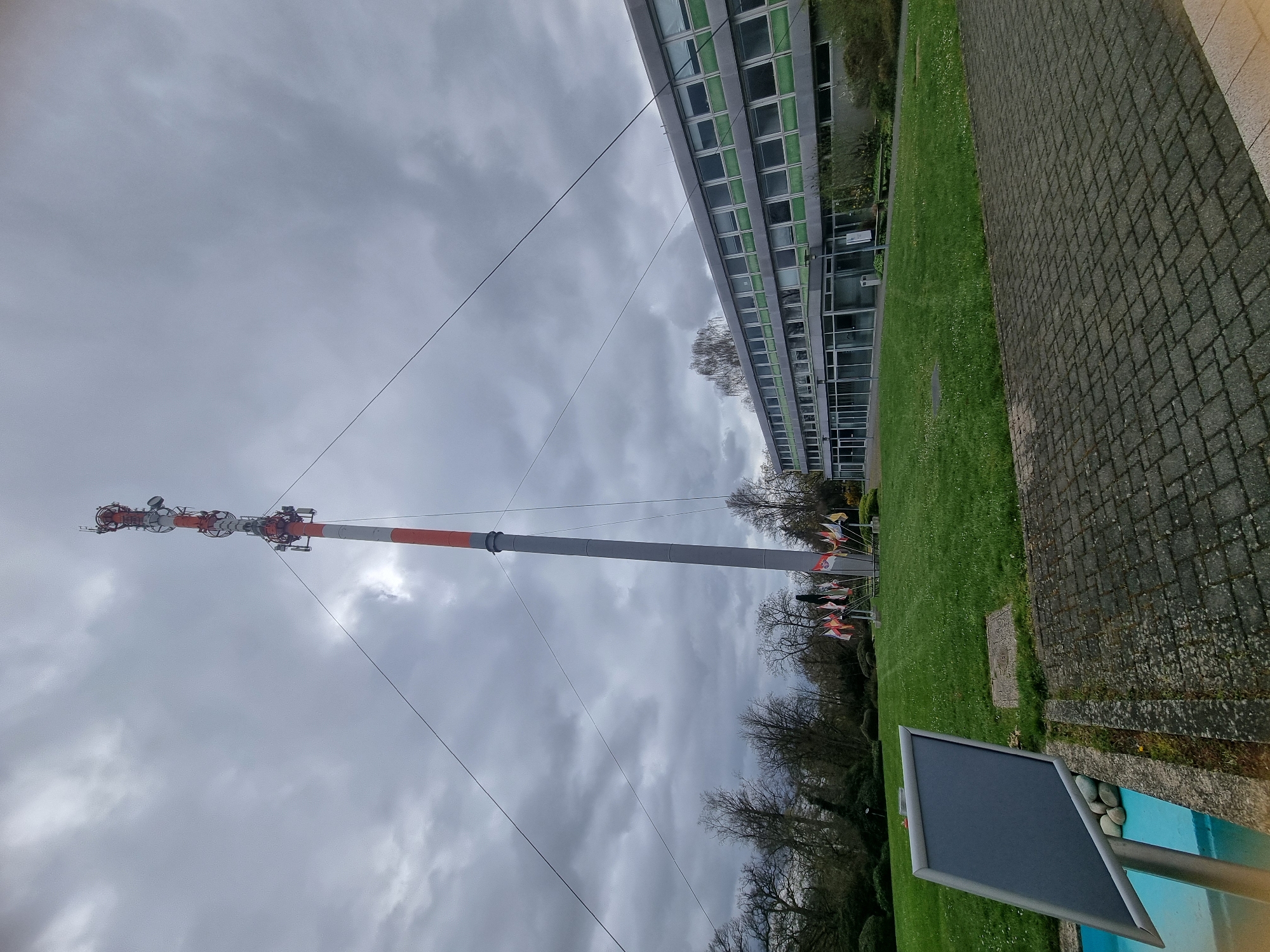 The image depicts the radio tower in Saarbrücken, Germany, on an overcast day. The tower, colored gray with red and white accents near the top, dominates the view. It is surrounded by a green grassy area, a building with multiple windows, and a brick walkway. In the foreground, there is a sign with a dark surface. Bare trees are visible in the background. Several wires extend from the top of the tower toward the ground. There is no one visible in the image.