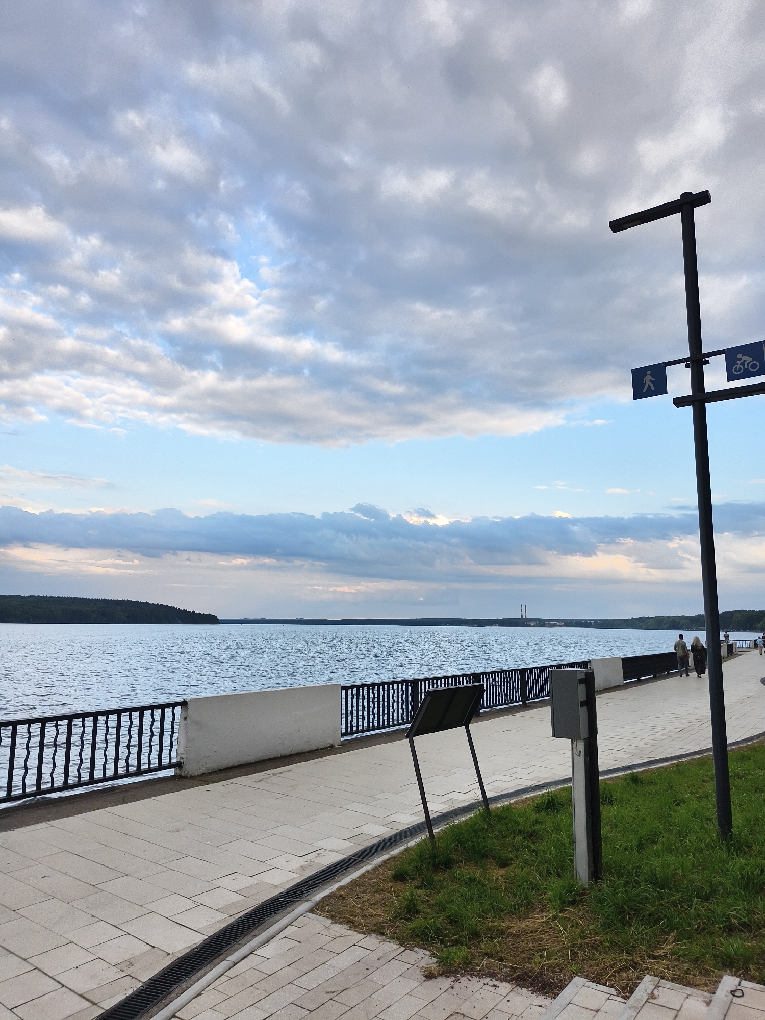 This is an outdoor, eye-level, vertical photograph of a waterfront promenade. The image captures a wide expanse of calm, blue water to the left, bordered by a treeline on the distant horizon. The sky above is filled with a mix of white and gray clouds, with patches of blue sky visible.
In the foreground, a light-colored paved promenade runs horizontally across the image. A black metal railing with vertical bars separates the promenade from the water. Behind the railing, a white concrete wall is visible. Further to the right on the promenade, a black lamppost stands, with two blue directional signs attached to it: one with a pedestrian silhouette and another with a bicycle silhouette. Two people are walking away from the camera on the promenade. A green grassy area with some dry grass is present to the right of the promenade, with a grey electrical box mounted on a pole. A black metal grate is installed along the edge of the promenade. In the distance, industrial smokestacks are visible on the horizon.
The scene depicts a public area for walking and recreation along a body of water, likely a river or lake, in the city of Chaikovsky, Russia. The time of day appears to be late afternoon or early evening, given the lighting and cloud cover.
