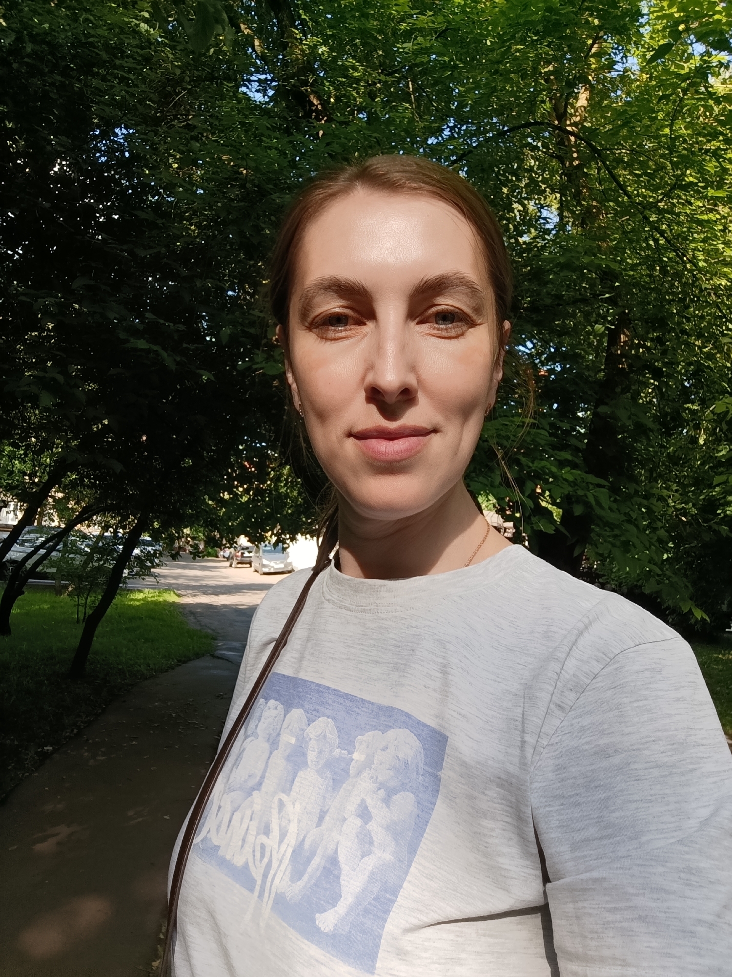 A woman, likely in her late 20s or 30s, is captured in a front-facing photograph, possibly a selfie, outdoors in Moscow, Russia. She has light brown hair pulled back and looks directly at the camera with a calm, pleasant expression and a slight smile. She is wearing a light grey marled t-shirt with a light blue graphic print on the front depicting several abstract or classical-style figures. A thin, dark strap from a bag or accessory crosses her chest, and a delicate gold-colored necklace is visible.
The setting is an urban or suburban green space, possibly a park or tree-lined street, on a bright, sunny day. The background is filled with dense, lush green foliage of trees, through which dappled sunlight filters, casting shadows and highlights. A paved pathway or sidewalk is visible to the woman's left, and in the further background, parked cars can be seen along a street, suggesting a residential or public area. The strong sunlight and vibrant green leaves indicate it is a warm season, likely late spring or summer. No legible text is visible on her shirt graphic.