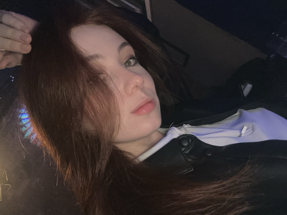 A person with auburn hair and light eyes gazes upward. They are wearing a black leather jacket over a white and light blue top. A hand appears to be touching their hair on the left side. The background is dark, with a faint, colorful, abstract shape visible to the left of the person. The individual's expression is calm and serene.