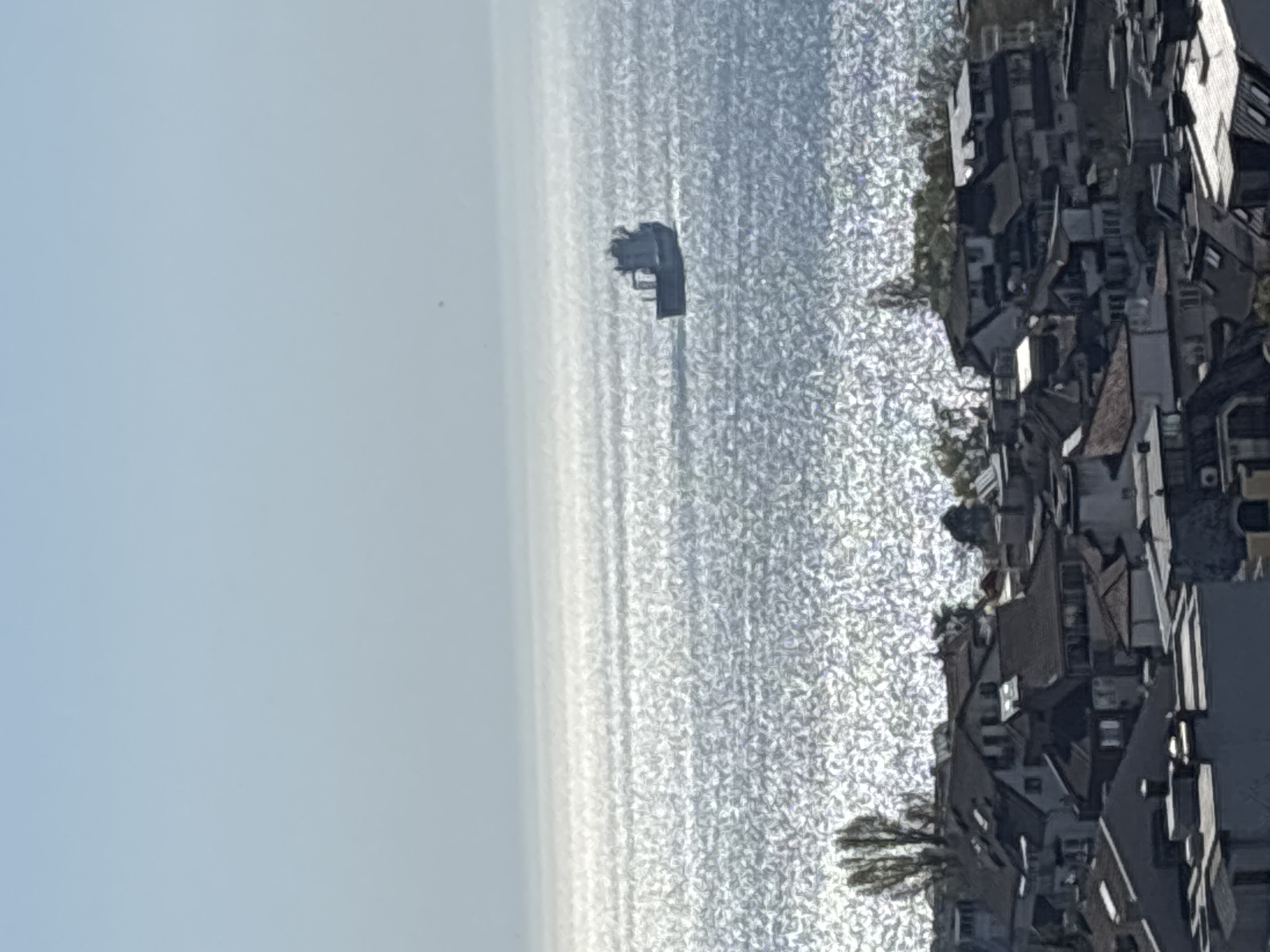 The image shows a body of water with a ship in the distance. On the right side of the image are densely packed buildings, possibly a residential area. The sky is clear and light blue. The location is likely near a coastal city in Unknown, Unknown.