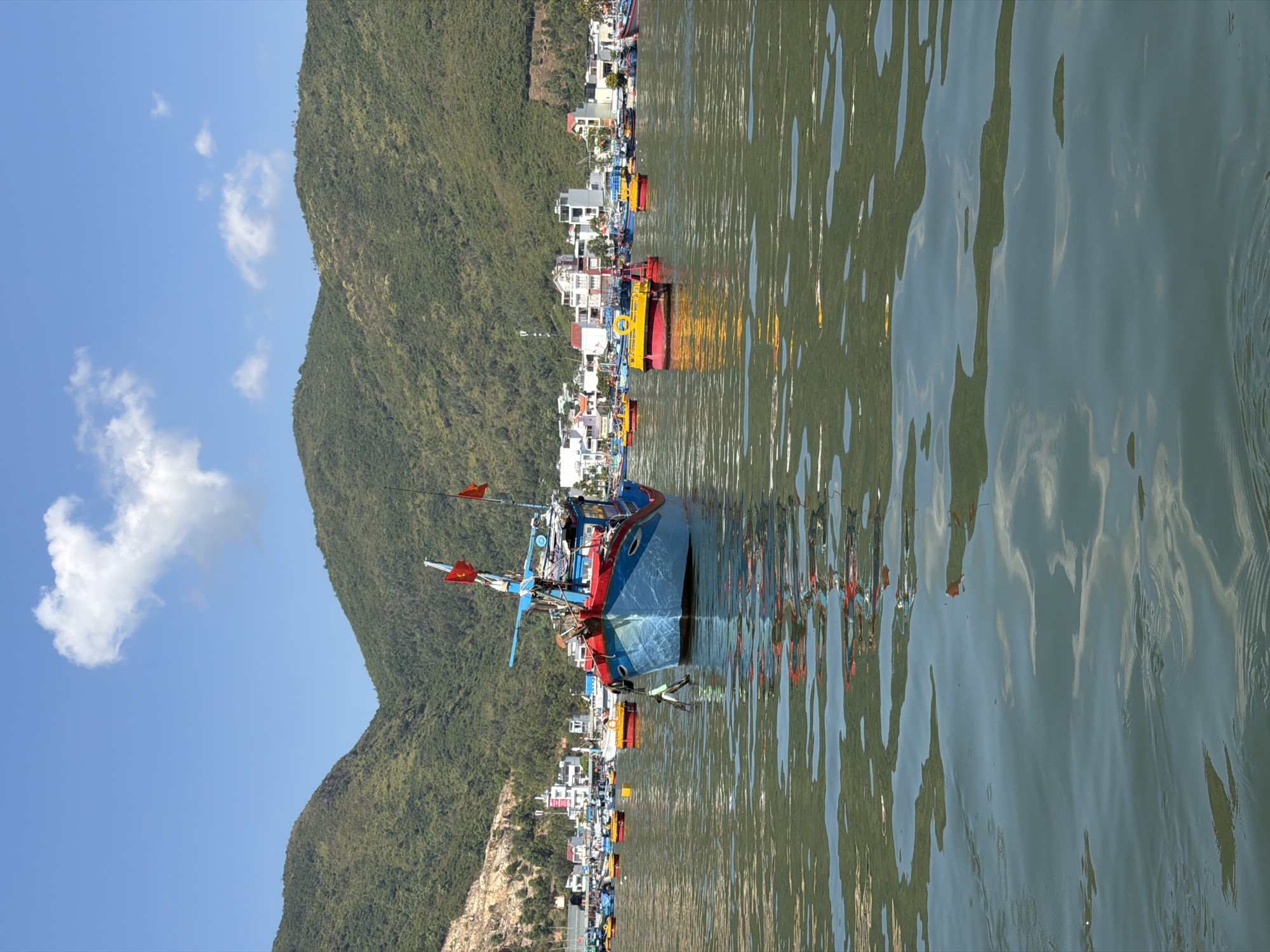 The image depicts a coastal scene, likely in Nam Nha Trang Ward, Vietnam. A body of water, possibly a bay or inlet, dominates the right side of the frame, with visible ripples on the surface reflecting the sky, mountain, and boats. Several fishing boats are docked along the shoreline, with a prominent blue and red boat closer to the foreground. Behind the boats, there is a coastal town with tightly packed buildings, which appear to be mostly white or light-colored. Behind the buildings, a large, green mountain rises. The sky is blue with scattered white clouds on the left, contrasting the green mountain.